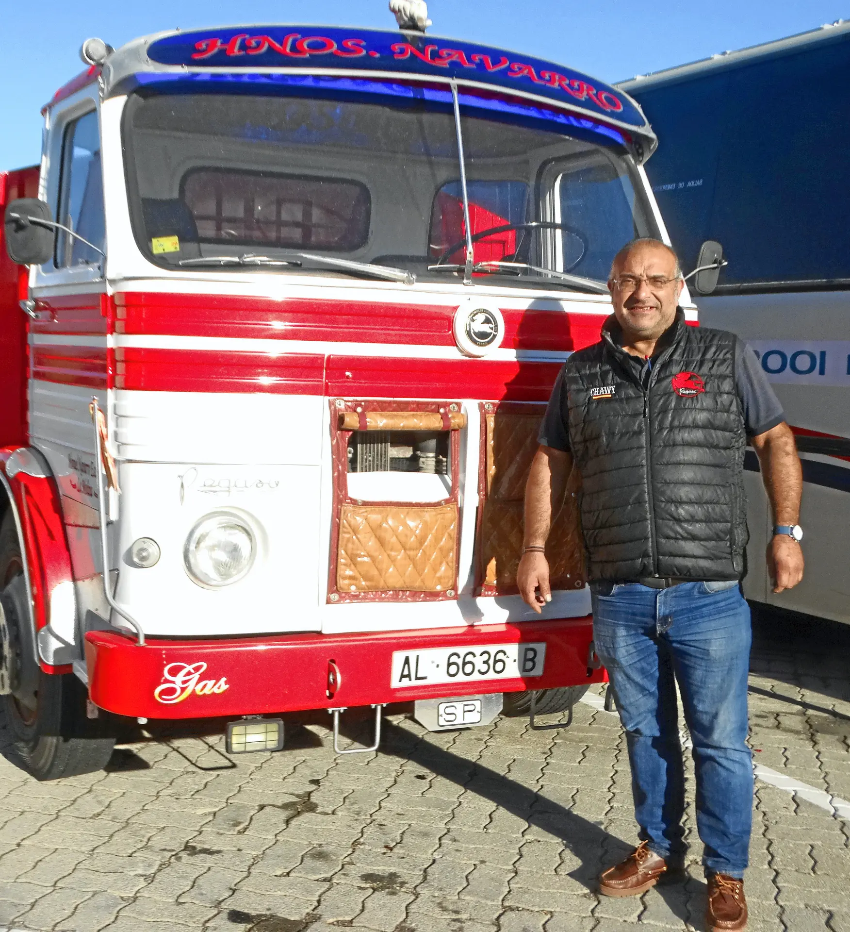 Un hombre sonríe delante de un camión rojo y blanco de época con la inscripción "Hnos. Navarro" y una pegatina de "Borrador automático" en el parabrisas.