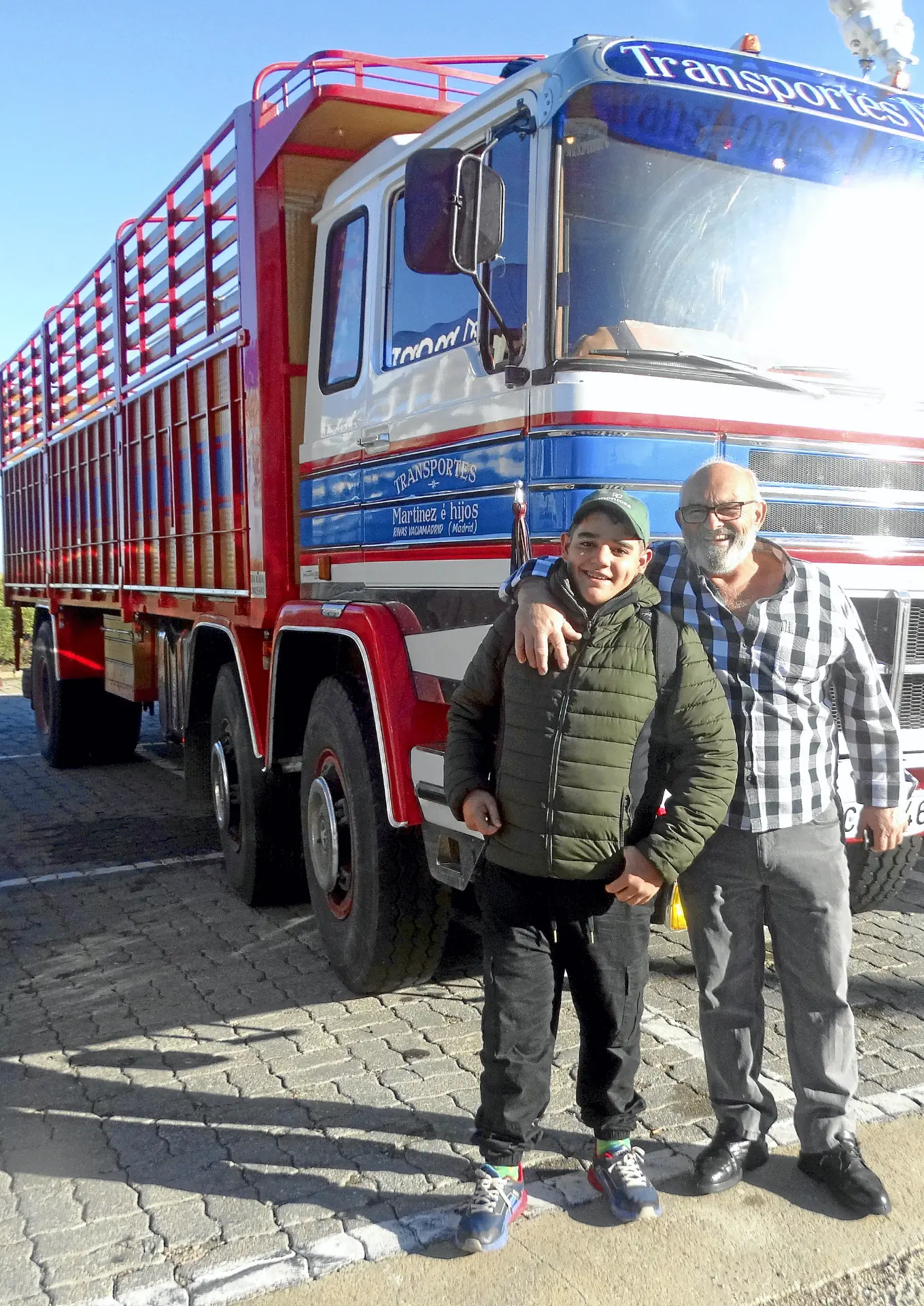 Dos hombres sonríen y posan delante de un gran camión de transporte rojo y azul, uno de los camiones clásicos que se ven a lo largo de la Ribera del Duero, en un día soleado.