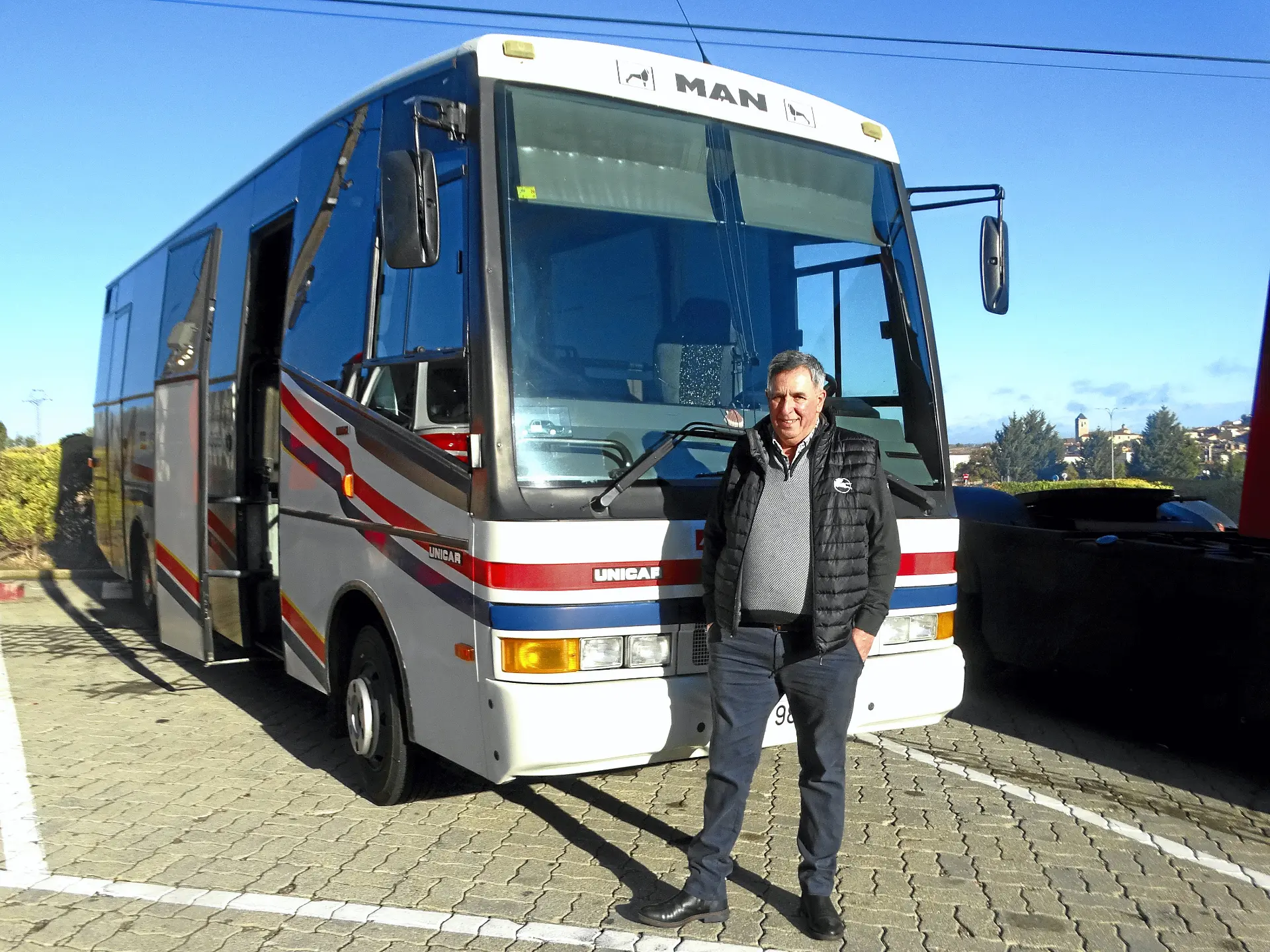 Un hombre se para frente a un autobús aparcado con franjas rojas y azules en un día soleado, listo para iniciar su ruta por la Ribera del Duero, por donde suelen pasar camiones clásicos.