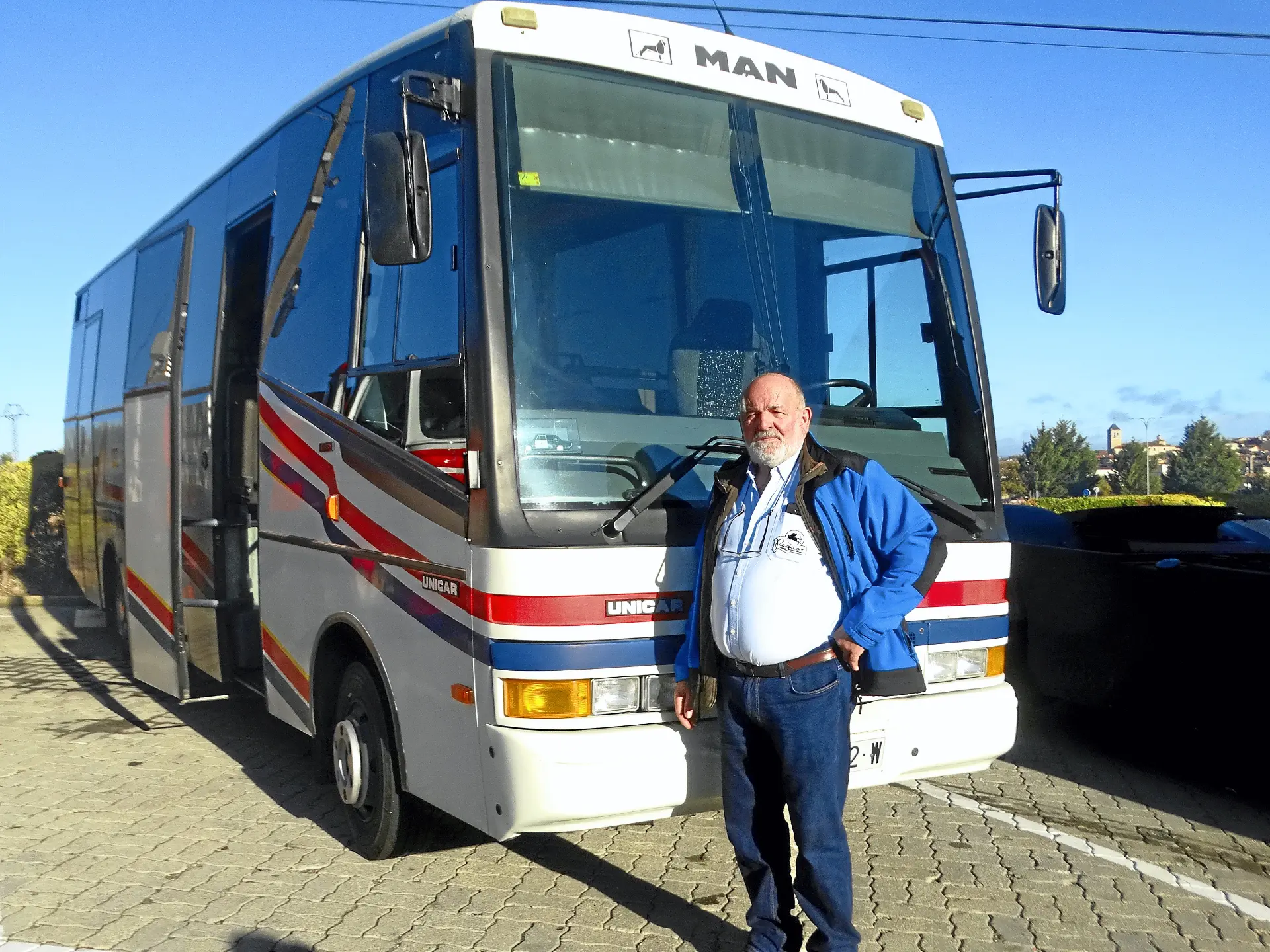 Un hombre mayor con chaqueta azul se para delante de un autobús blanco aparcado con rayas rojas y amarillas, que recuerda a los camiones clásicos que se ven por la Ribera del Duero.