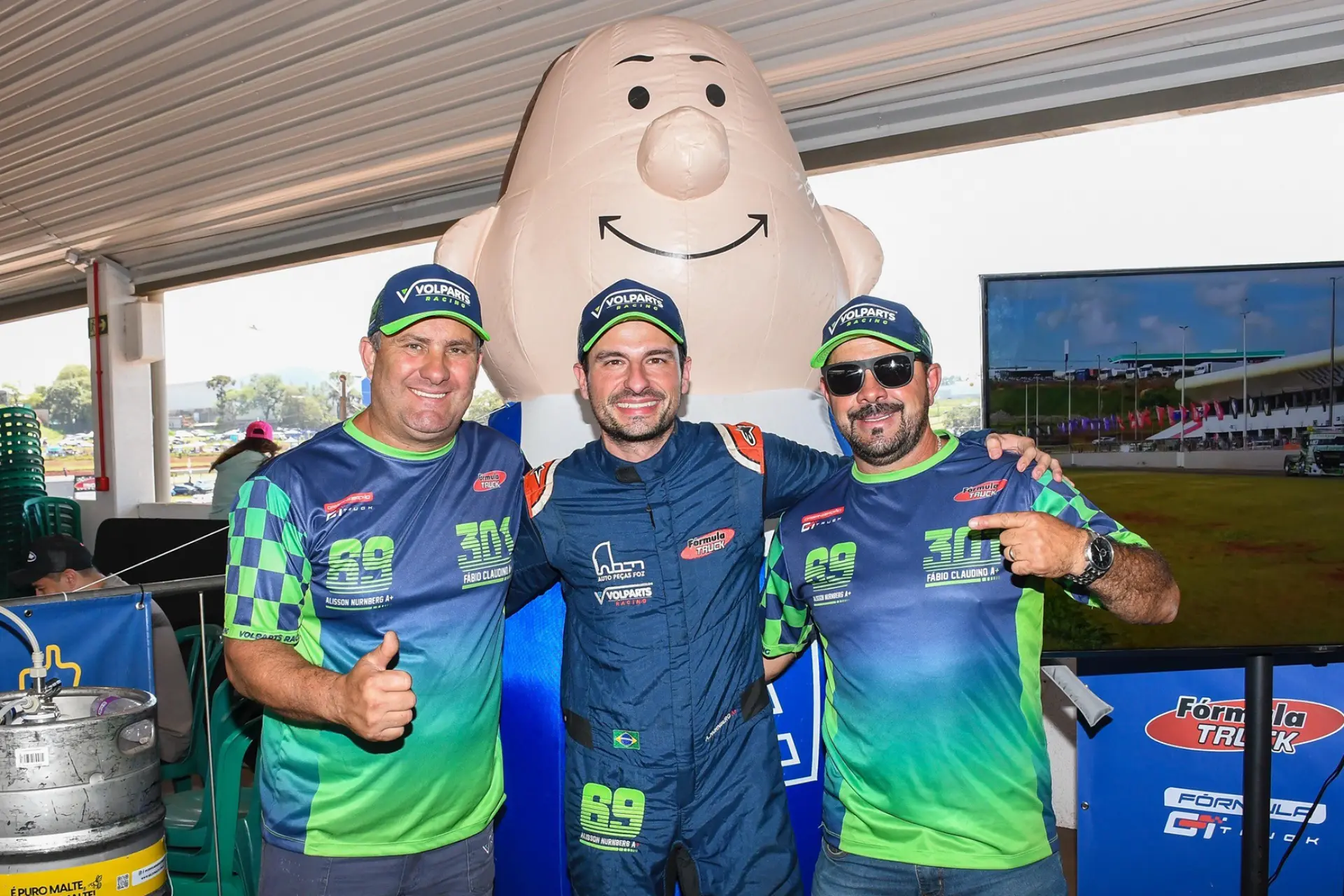 Tres hombres sonrientes posan juntos en un interior, uno de ellos con un mono de carreras, con un gran globo de dibujos animados y un cartel que dice "borrador automático" detrás de ellos.