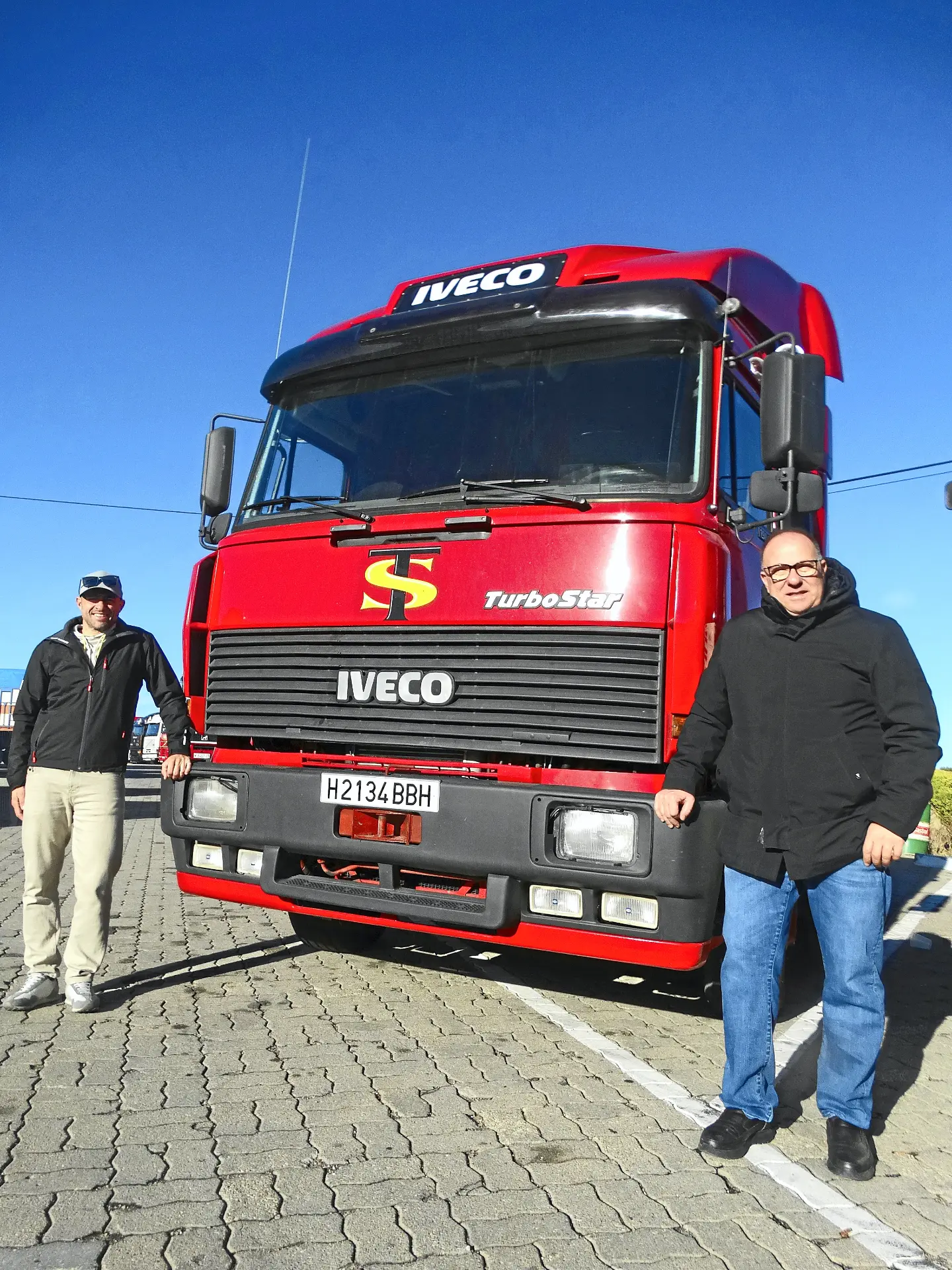 Dos hombres sonríen delante de un gran Iveco TurboStar rojo, uno de los camiones clásicos admirados por los aficionados, en un día soleado en la Ribera del Duero.