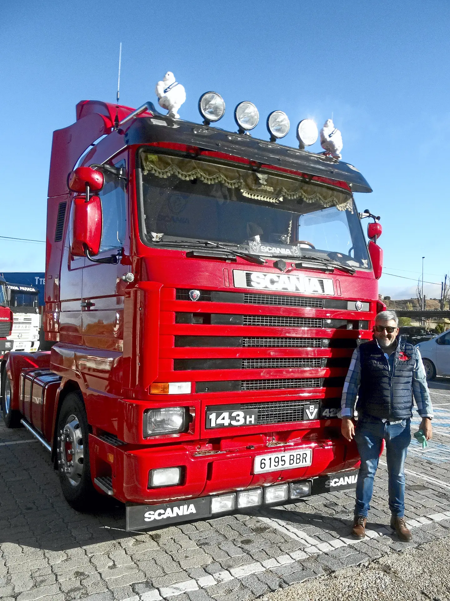 Un hombre junto a un gran camión Scania rojo aparcado en un día soleado, listo para continuar su ruta entre camiones clásicos.
