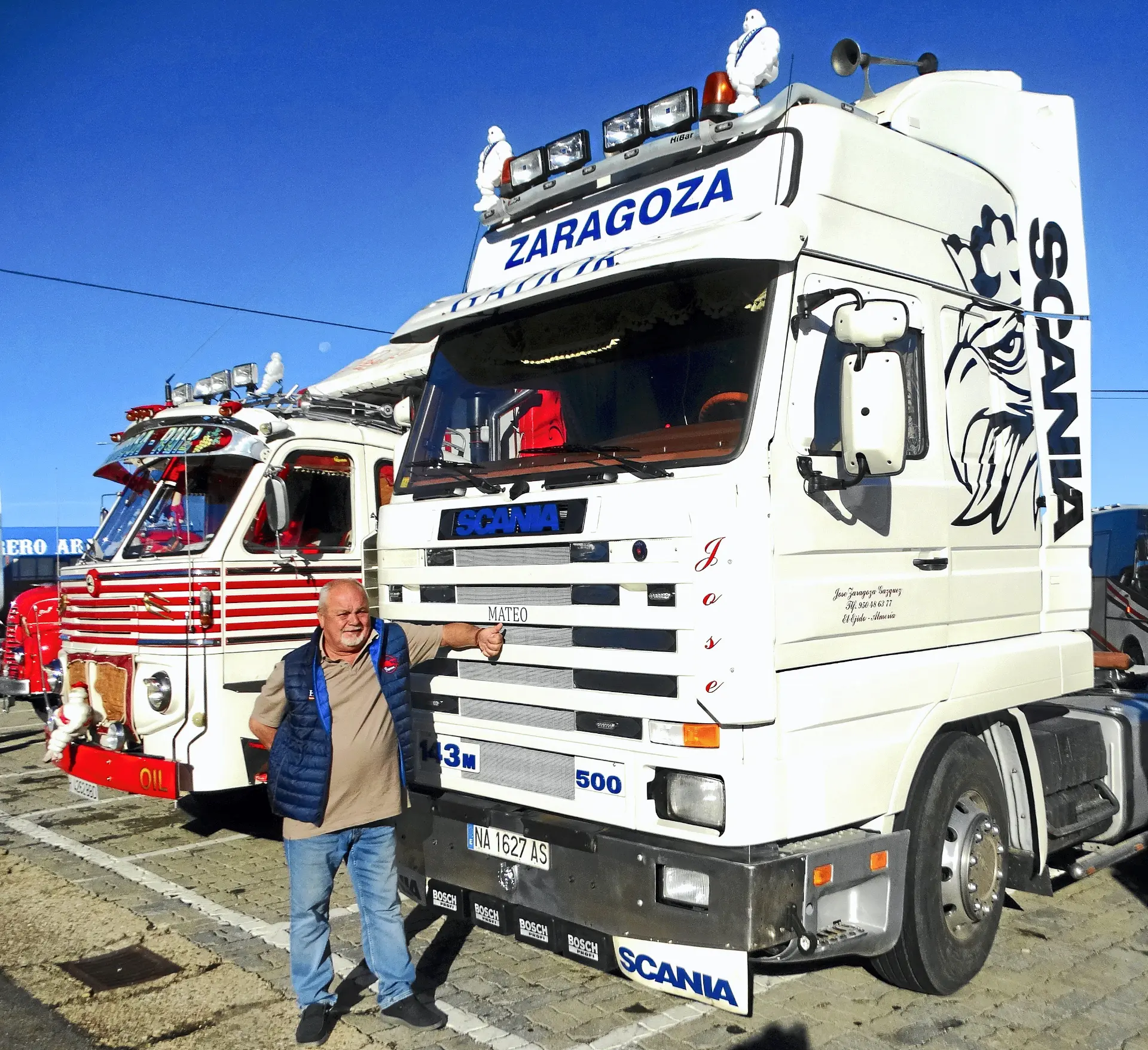 Un hombre sonríe junto a un camión Scania blanco rotulado "Zaragoza", con camiones clásicos y un camión rojo aparcados al lado.