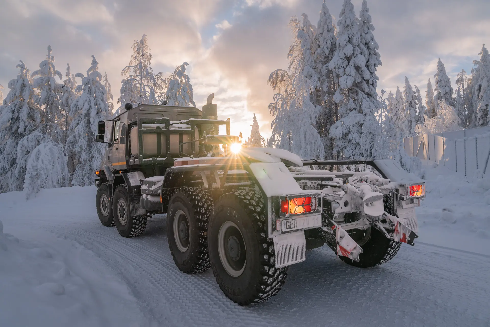 Un Mercedes-Benz Zetros circula por una carretera nevada a través de un bosque de árboles cubiertos de nieve al amanecer o al atardecer, navegando por el paisaje de Finlandia durante las pruebas árticas.