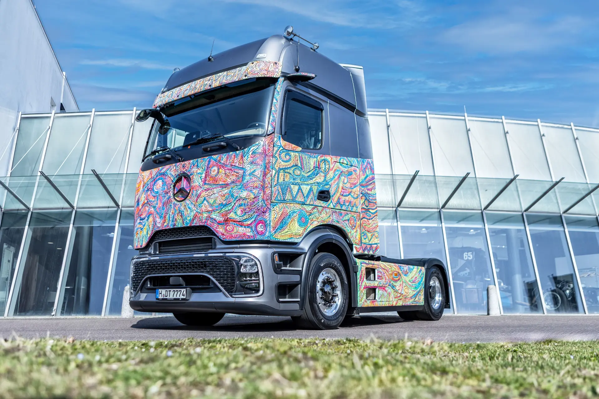 Un Mercedes-Benz art truck edición Actros con coloridos motivos psicodélicos pintados en la cabina, aparcado frente a un moderno edificio, exhibiendo el estilo de los camiones de larga distancia.
