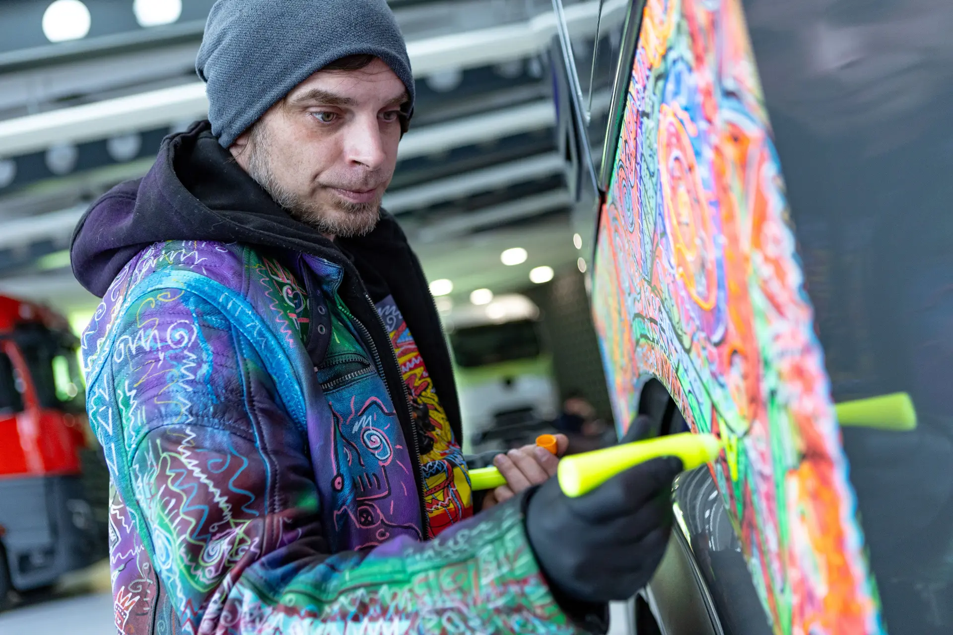 Un hombre con una colorida chaqueta pinta vibrantes diseños en el lateral de un Mercedes-Benz Actros L ProCabin, convirtiendo este camión decorado gira Actros Alemania en una conmovedora obra de arte utilizando herramientas amarillas.