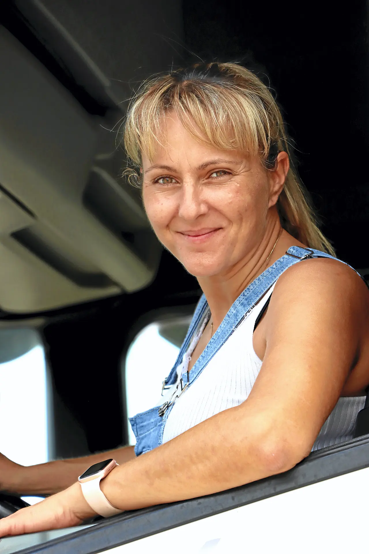 Una mujer con camiseta blanca de tirantes y mono vaquero sonríe sentada en el asiento del conductor de un vehículo, encarnando el espíritu de Soy Camionera.