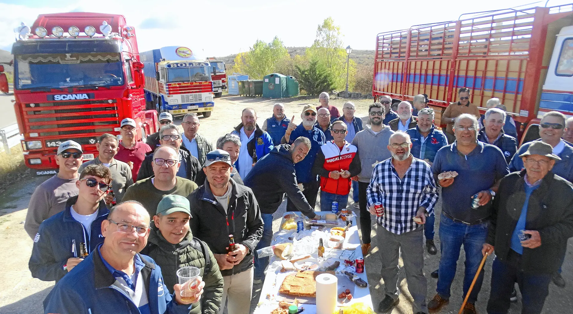 Un grupo de personas sonrientes y reunidas en torno a una mesa con comida y bebida cerca de varios camiones clásicos al aire libre.