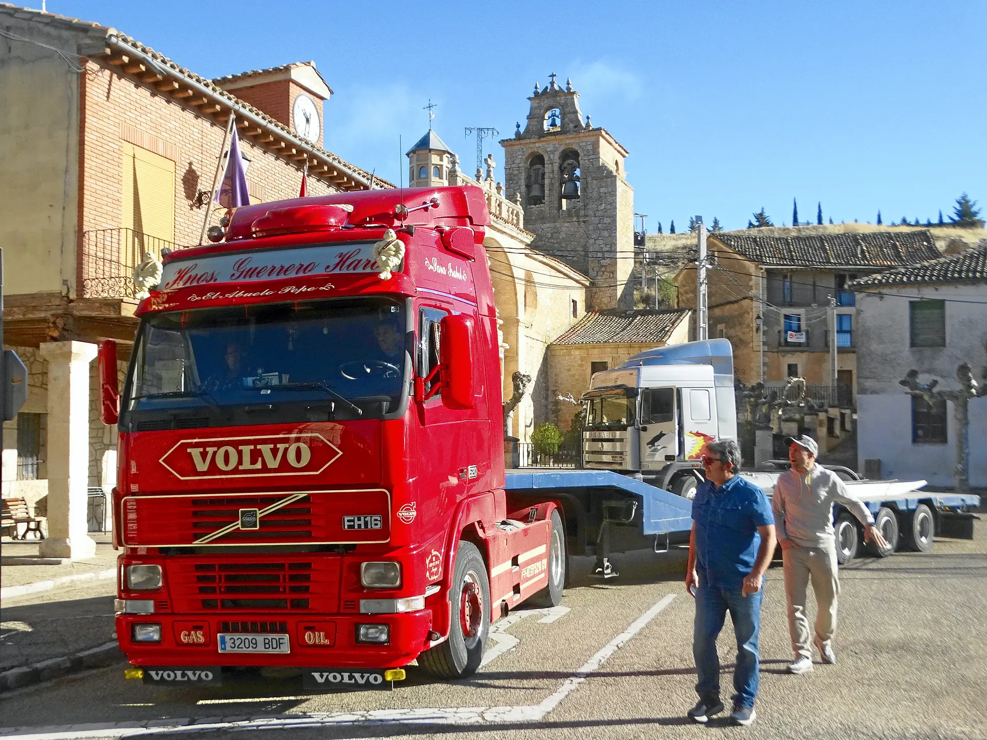 Un camión Volvo de color rojo brillante, que recuerda a los camiones clásicos, está aparcado en una calle con dos hombres caminando cerca en un entorno de pueblo europeo, evocando el encanto de la Ribera del Duero.