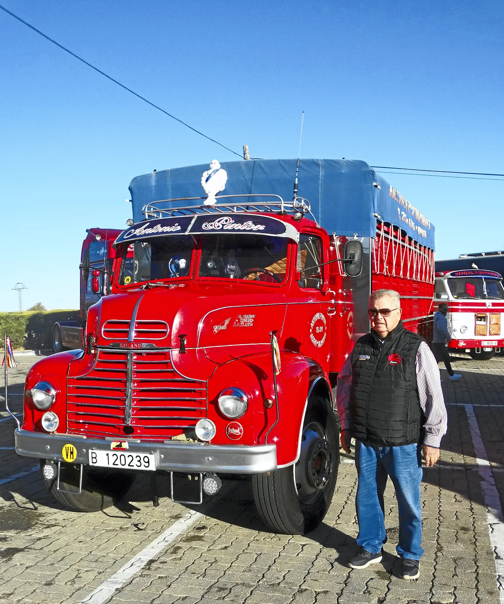 Un hombre junto a un camión rojo de época con la zona de carga cubierta de azul en un aparcamiento, destacando el encanto de los camiones clásicos en la región de Ribera del Duero.