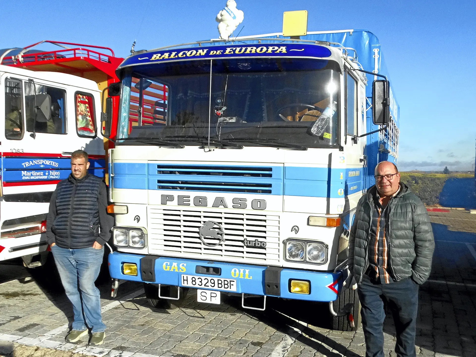 Dos hombres frente a un camión Pegaso azul con la etiqueta "BALCON DE EUROPA", aparcado junto a otro camión: una escena icónica para los amantes de los camiones clásicos, quizás vista a lo largo de la pintoresca Ribera del Duero.