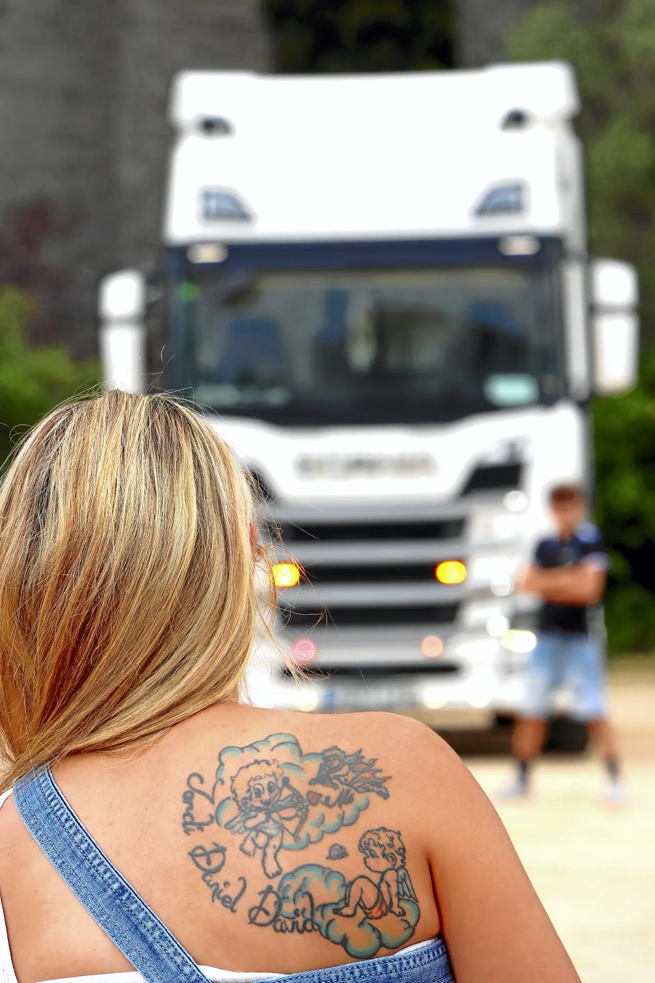 Una mujer con tatuajes en la espalda se enfrenta a un gran camión blanco, encarnando el espíritu de una auténtica camionera; un hombre aparece difuminado al fondo.