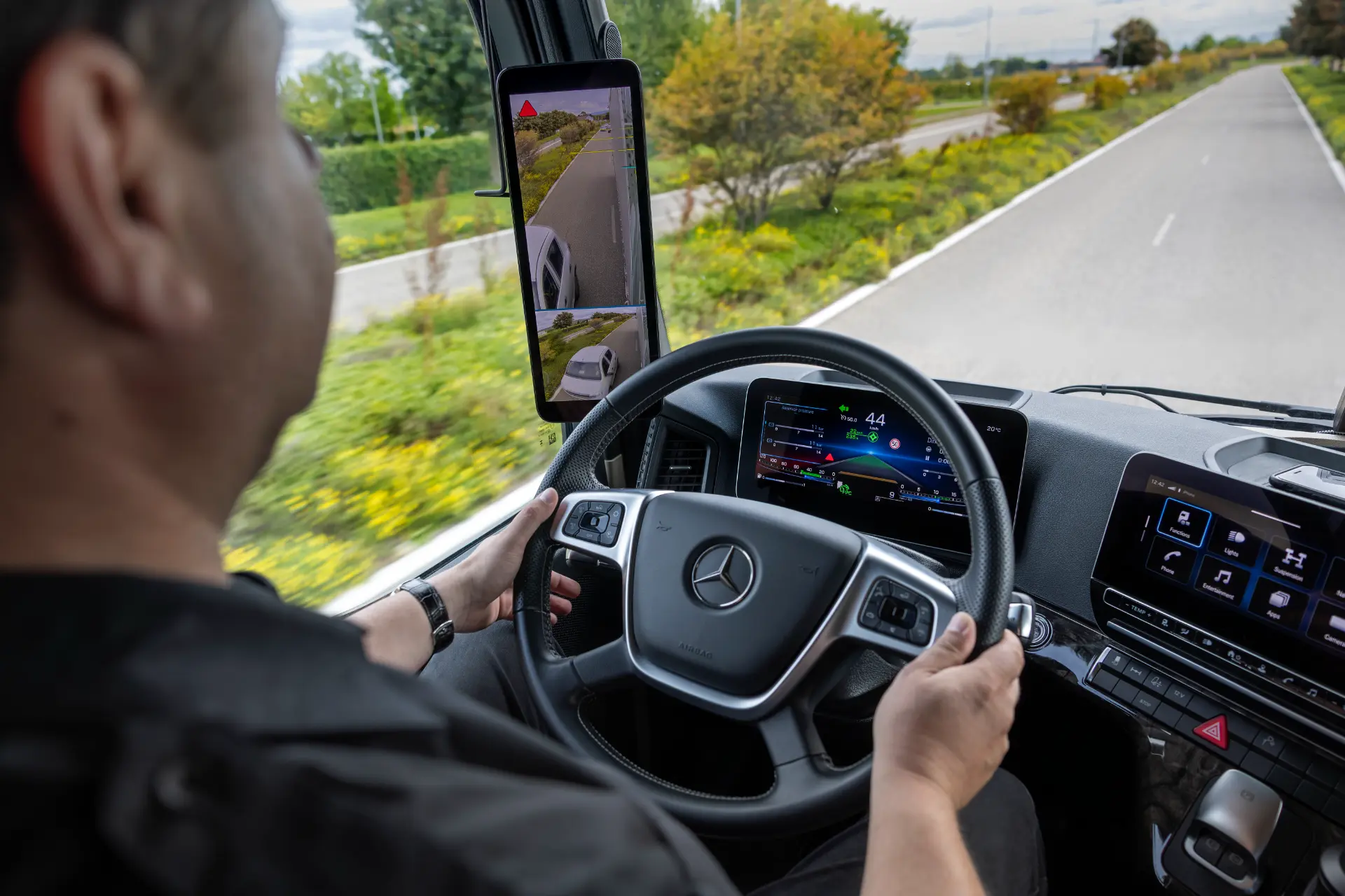 Persona conduciendo un camión Mercedes ProCabin, sujetando el volante, con pantallas digitales y un visor de cámara a la vista.