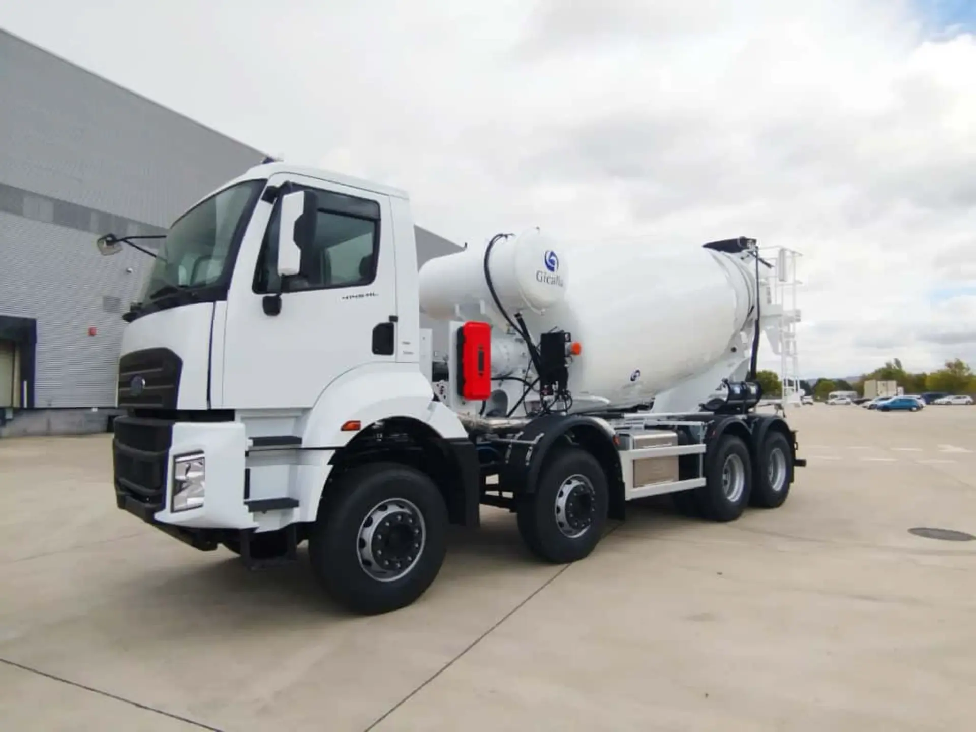 Camión hormigonera blanco aparcado en un solar de hormigón cerca de una gran nave industrial en un día nublado, que recuerda a los robustos camiones ford f-line expuestos en SMOPYC Zaragoza.