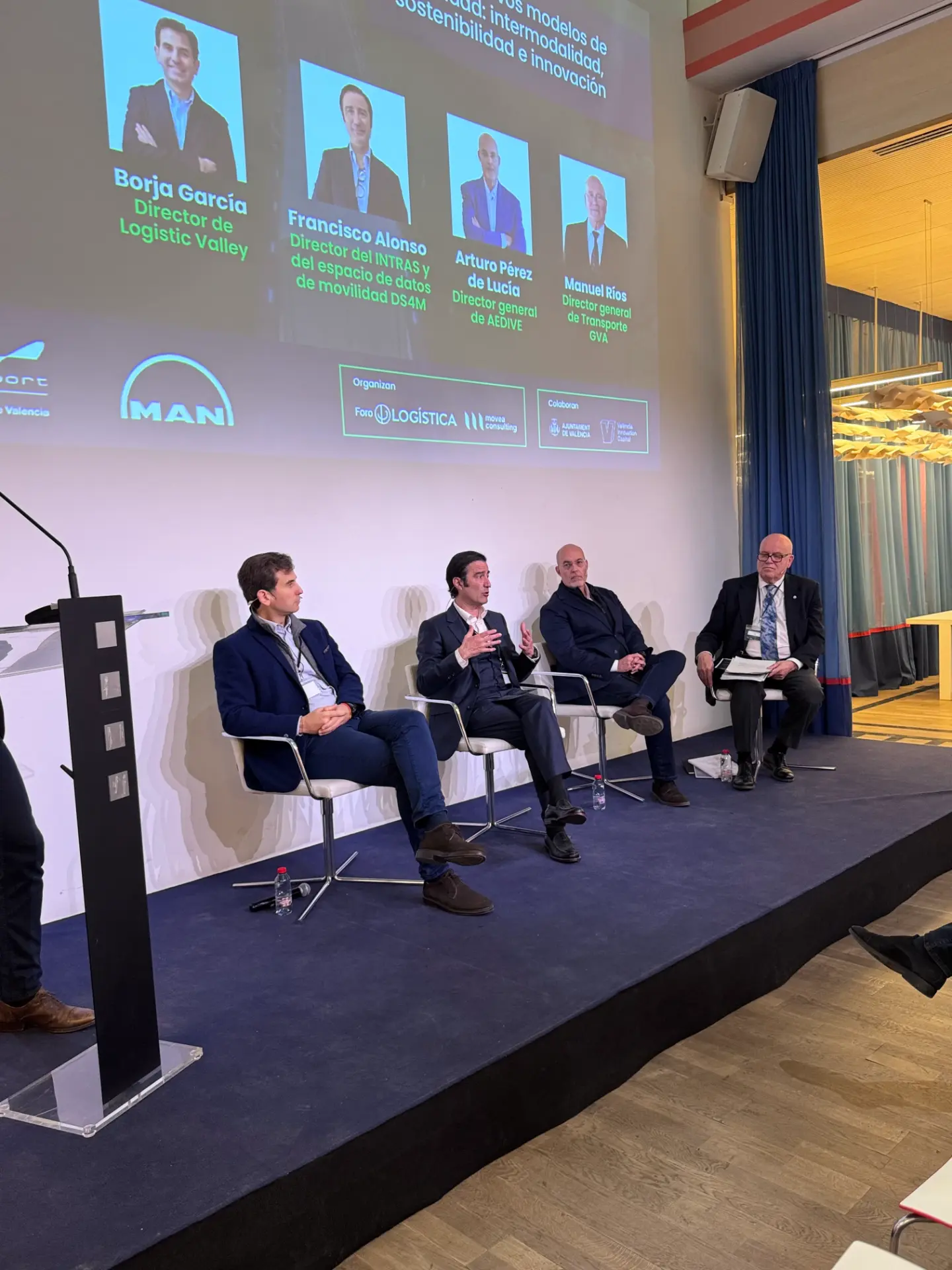 Cuatro hombres trajeados se sientan en el escenario de una mesa redonda en El Mobility Logistics Summit para hablar de logística y Movilidad Sostenible, mientras se muestran diapositivas de la presentación detrás de ellos.