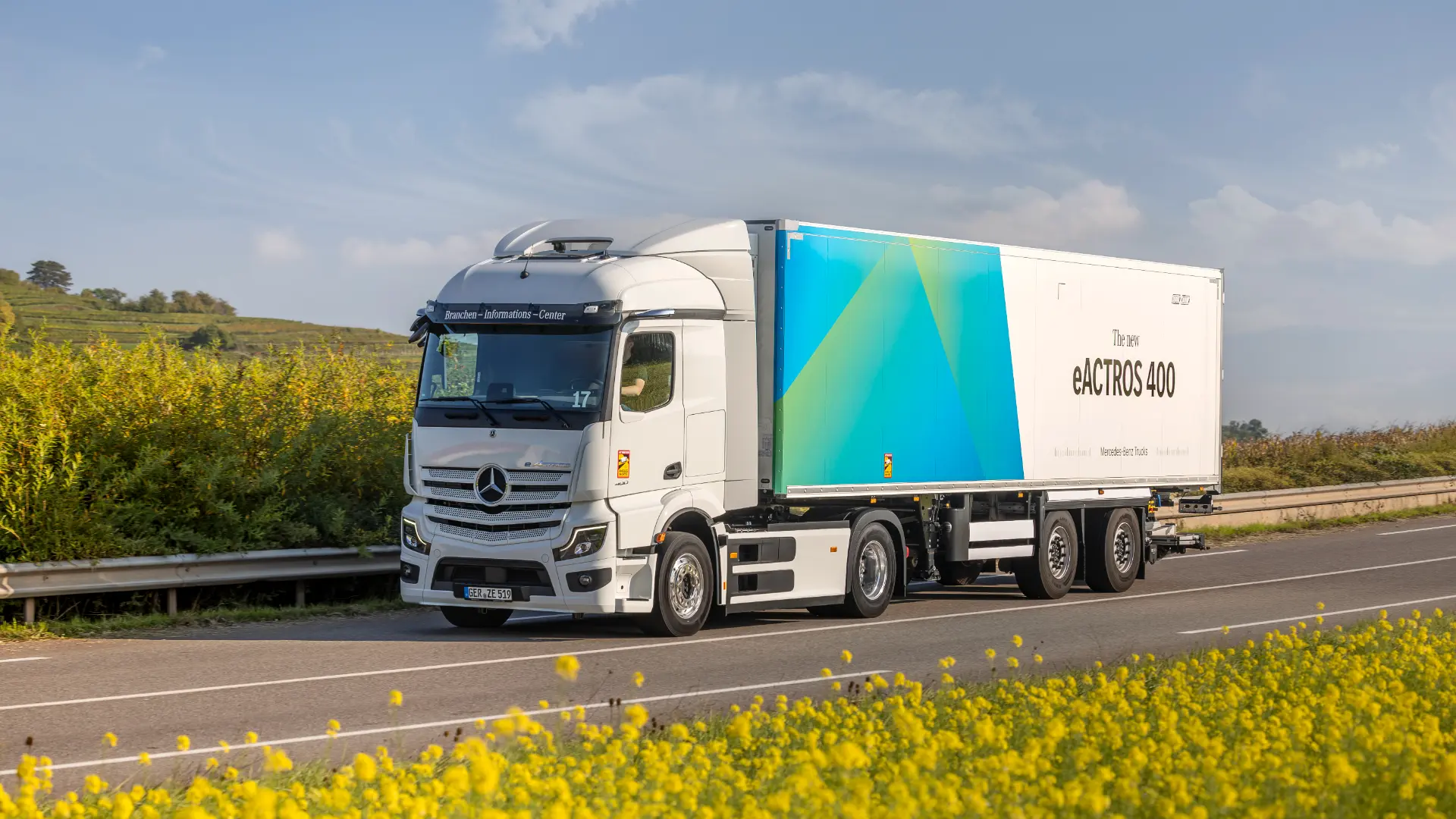 Un camión eléctrico Mercedes-Benz eActros 400 blanco con cabina Mercedes ProCabin circula por una carretera junto a un campo con flores amarillas.