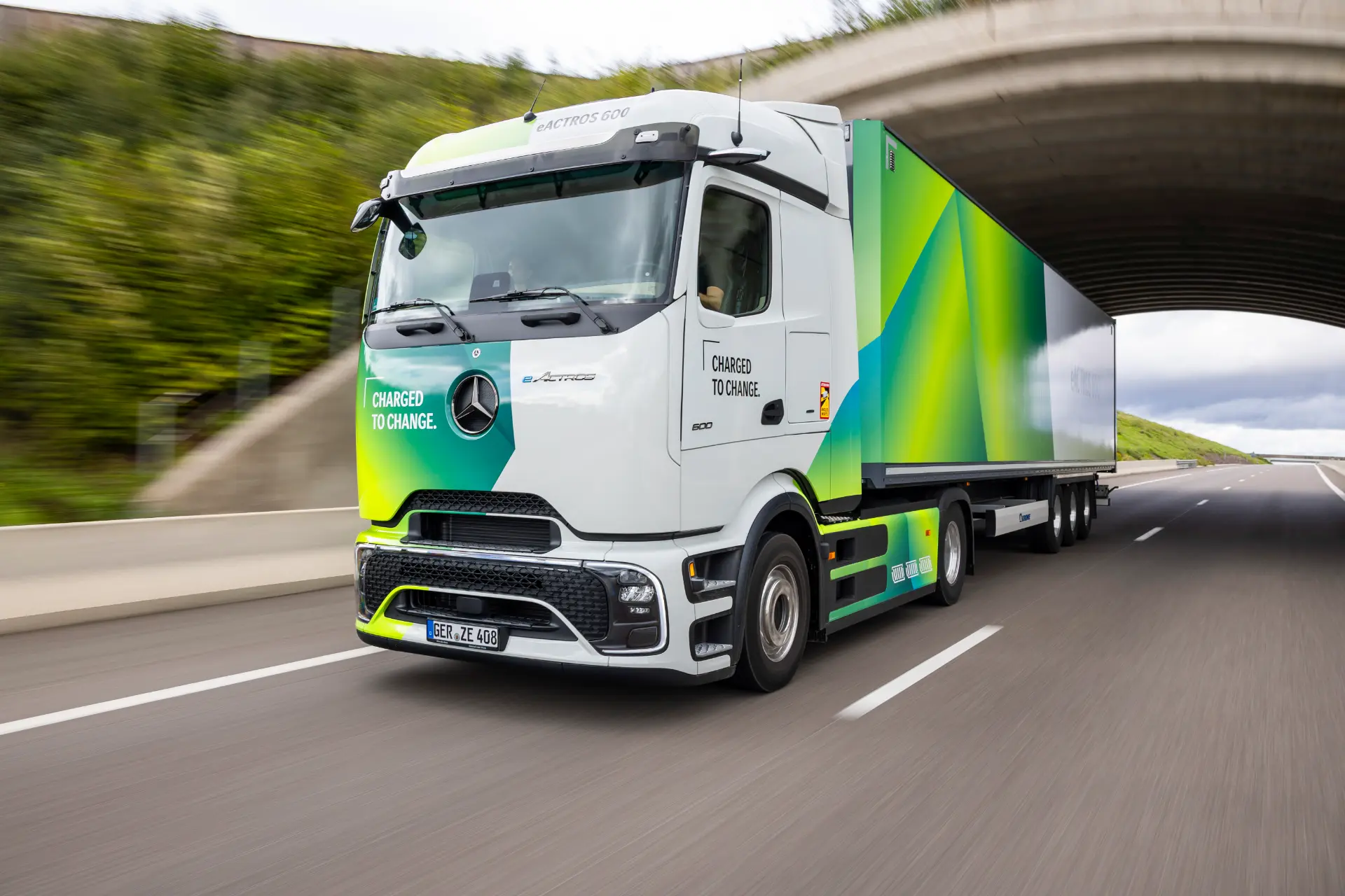Un semirremolque eléctrico Mercedes-Benz eActros 600 circula por una autopista bajo un paso elevado, arrastrando un colorido remolque.