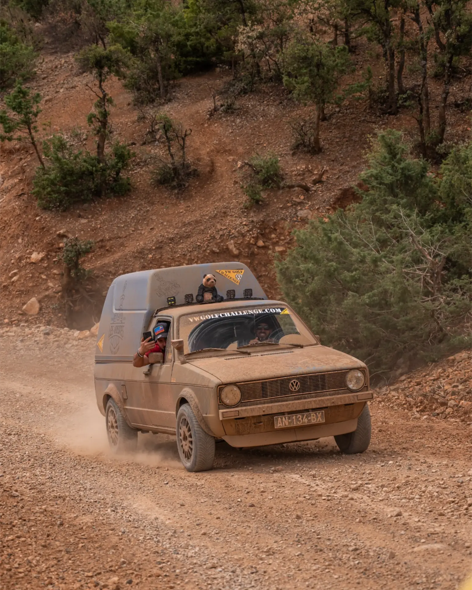 Un coche Volkswagen polvoriento circula por un camino de tierra rocosa con dos personas sonrientes en su interior, rodeado de árboles, durante la 12 edición Golf Challenge Algeciras.