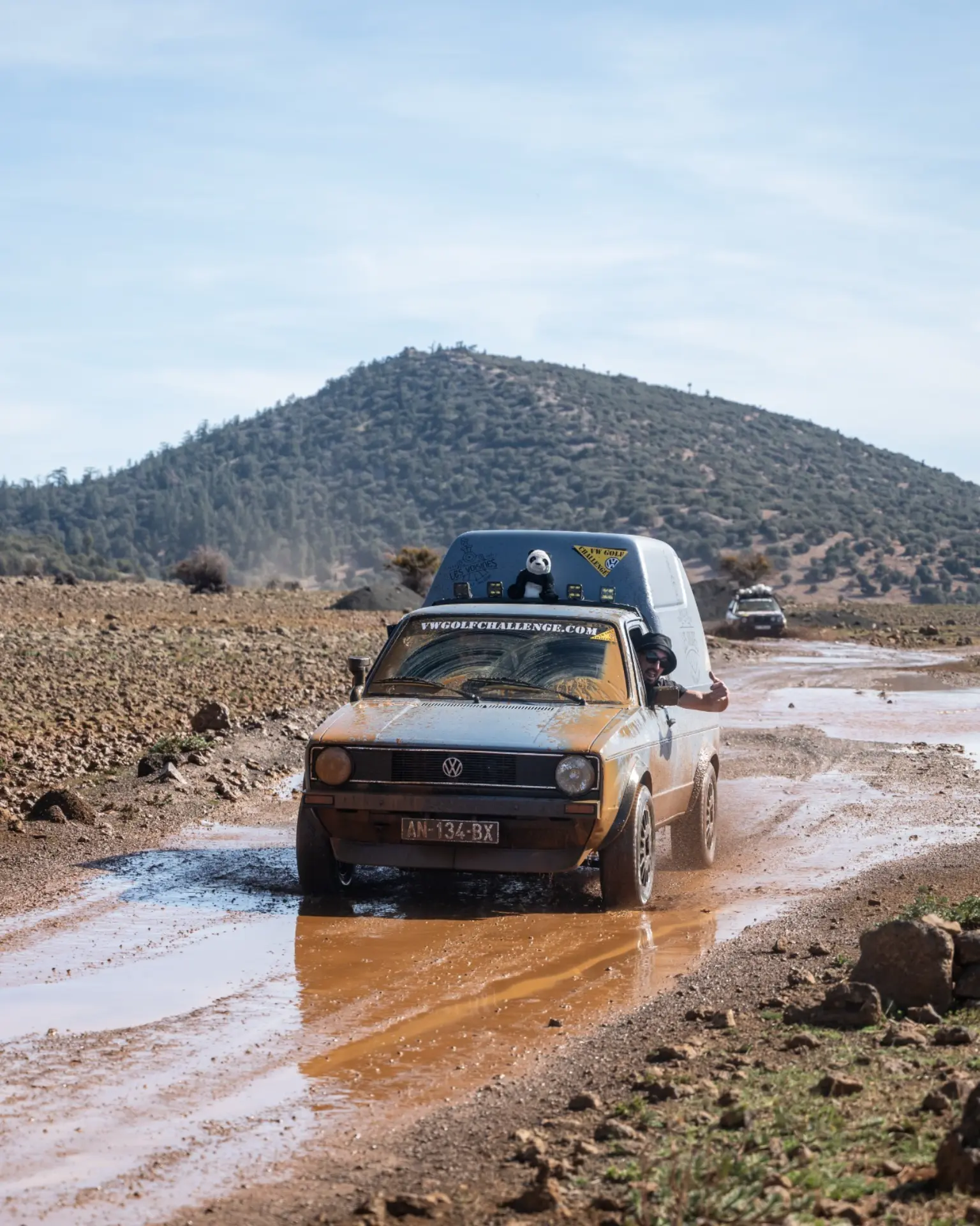 Un coche embarrado circula por una carretera mojada y pedregosa con montañas al fondo y otros vehículos detrás, capturando el espíritu aventurero de la 12 edición Golf Challenge Algeciras.