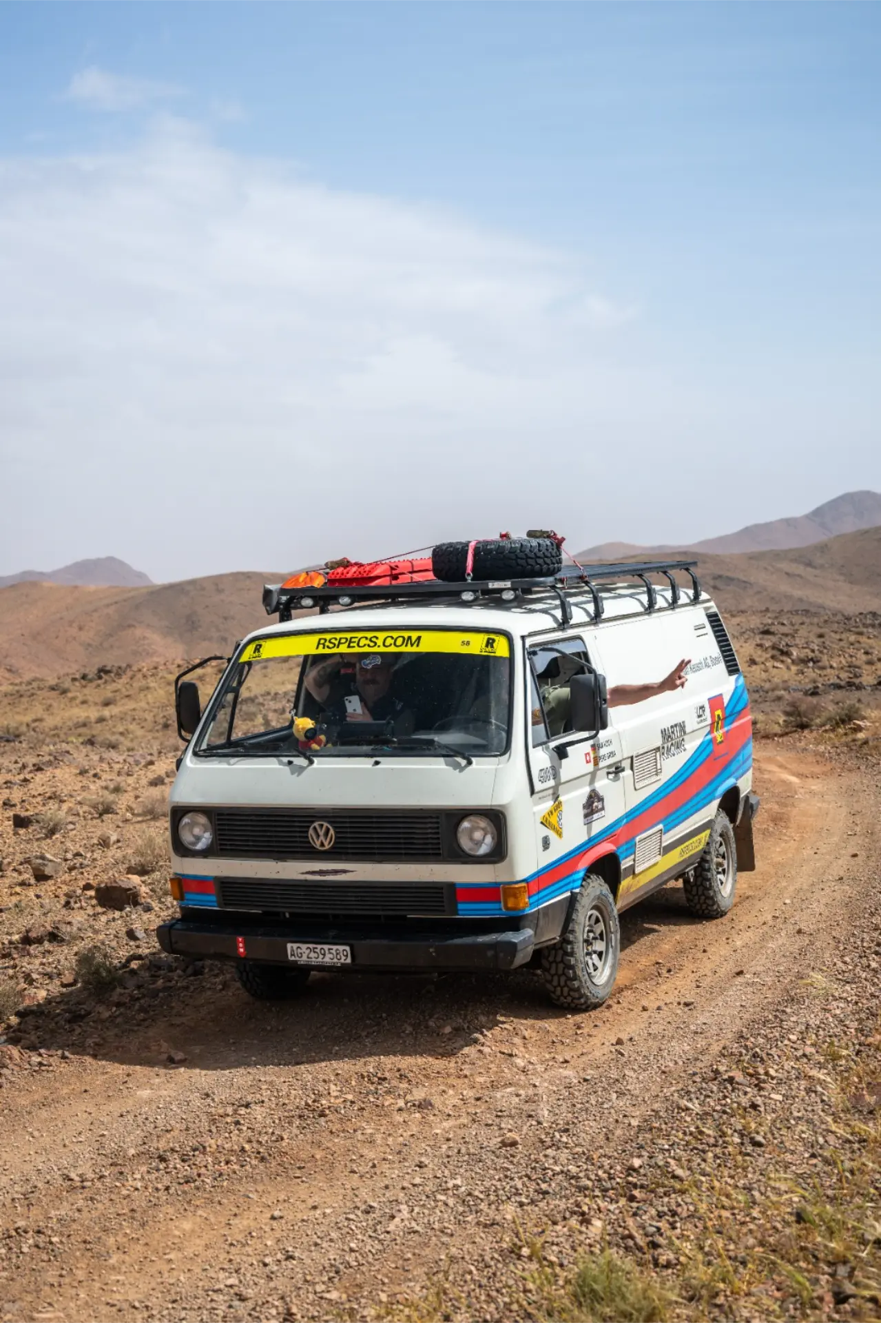 Una colorida furgoneta Volkswagen circula por una rocosa carretera desértica con montañas al fondo, capturando el espíritu aventurero del raid Volkswagen off-road pistas Marruecos.