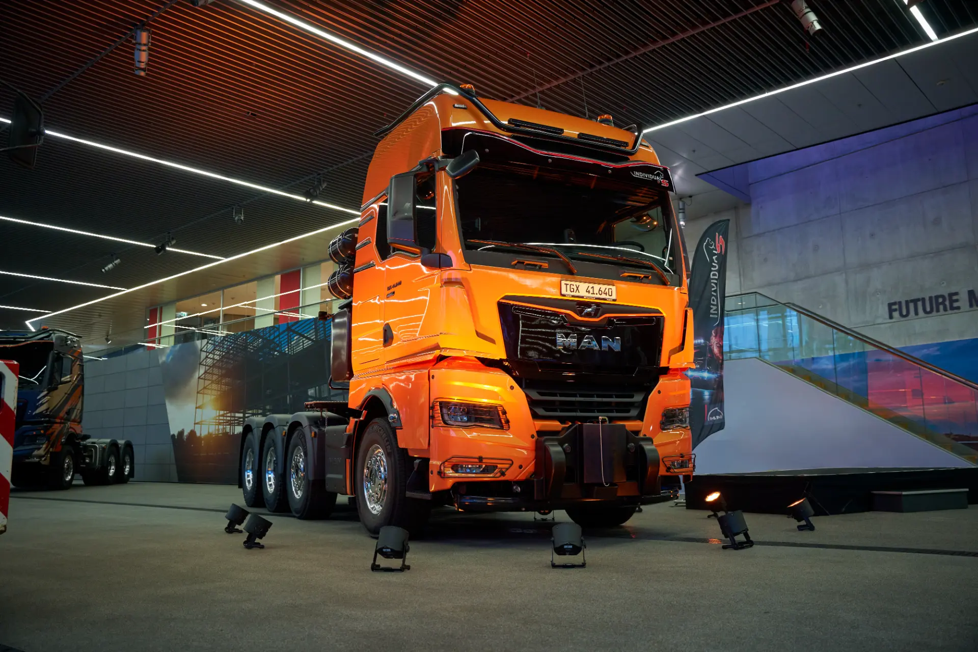 Una nueva tractora MAN naranja de 250 toneladas se exhibe en el interior, rodeada de una decoración moderna y focos, perfecta para la carga pesada de turbinas eólicas.