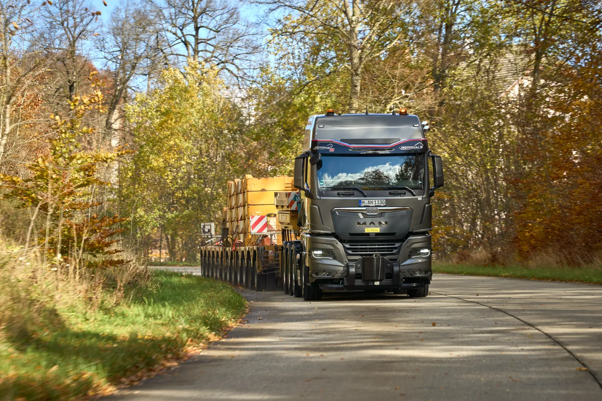 MAN TGX 41.640 negro de transporte pesado transportando una gran carga por una carretera sinuosa a través de una zona boscosa en otoño.