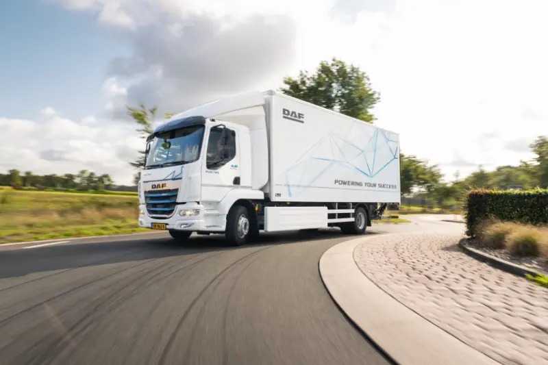 A white DAF electric truck, parte de las soluciones de carga para vehículos eléctricos, circula por una carretera curva rodeada de árboles y vegetación.