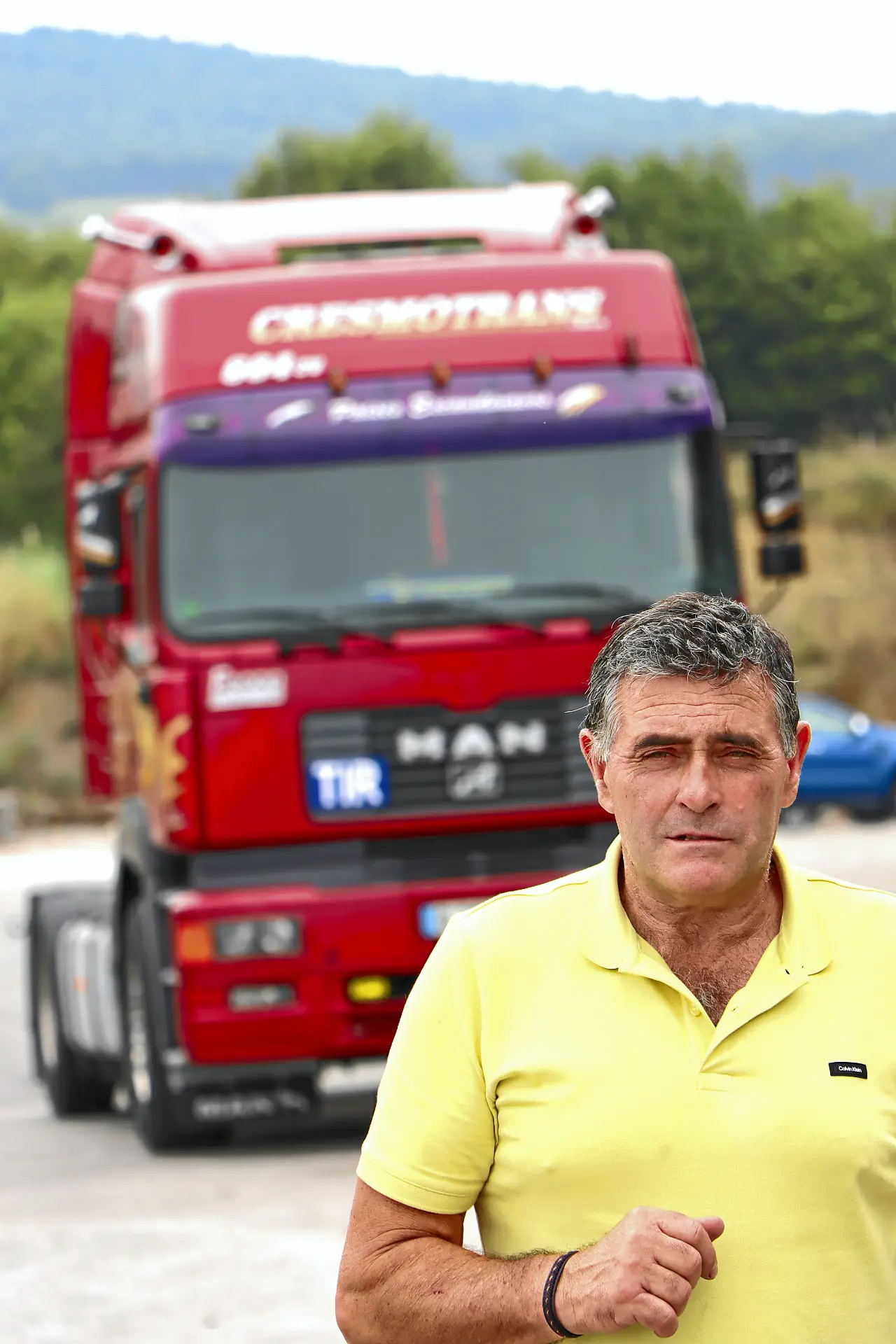 Hombre con camisa amarilla delante de un camión rojo Cresmotrans clásico aparcado en una carretera con árboles al fondo.