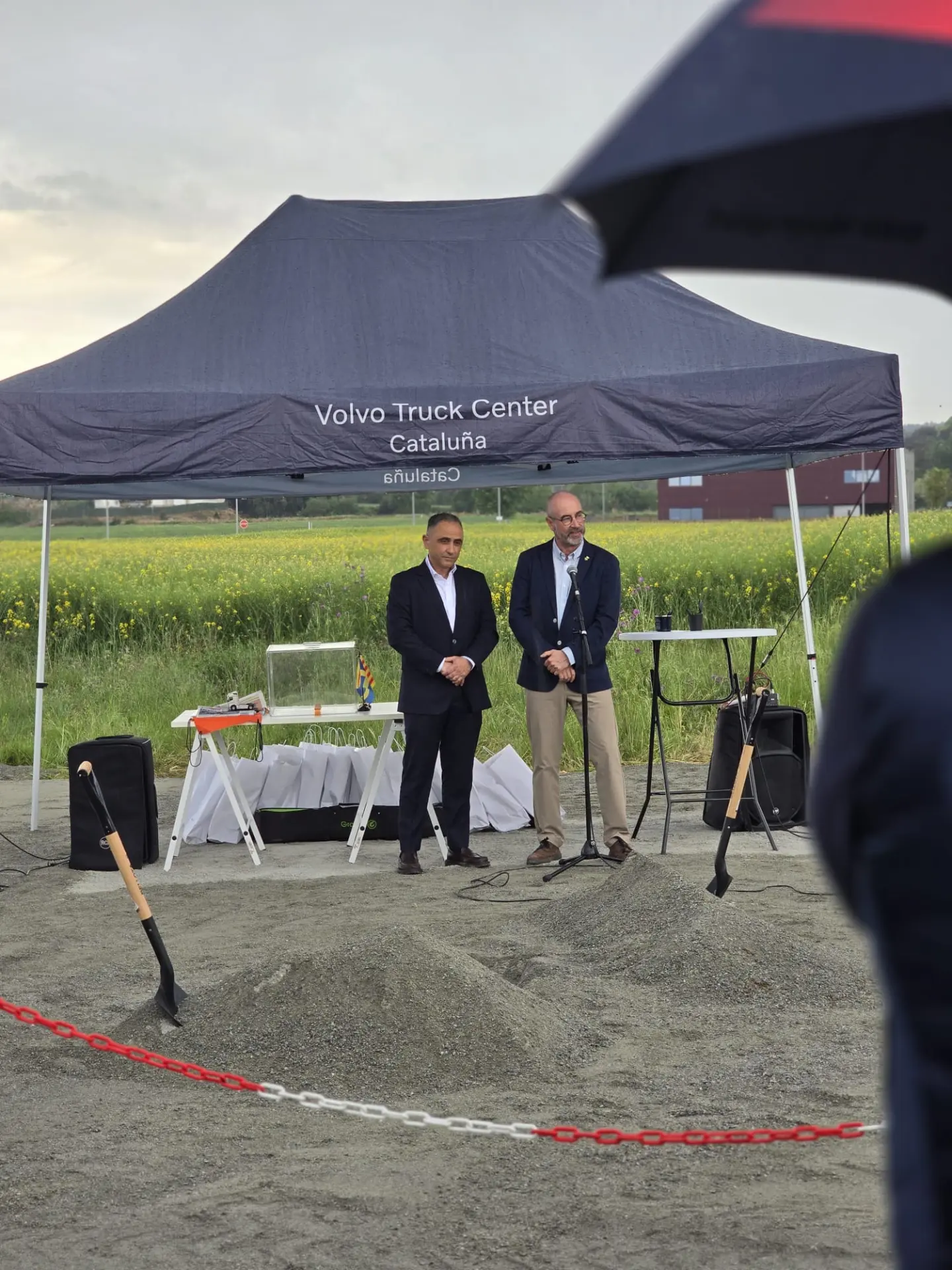 Dos hombres hablan bajo una carpa "Volvo Truck Center Cataluña" en un acto de colocación de la primera piedra con palas y una cinta roja, celebrando el futuro Truck Center Girona en Bescanó, un hito para Volvo Trucks.