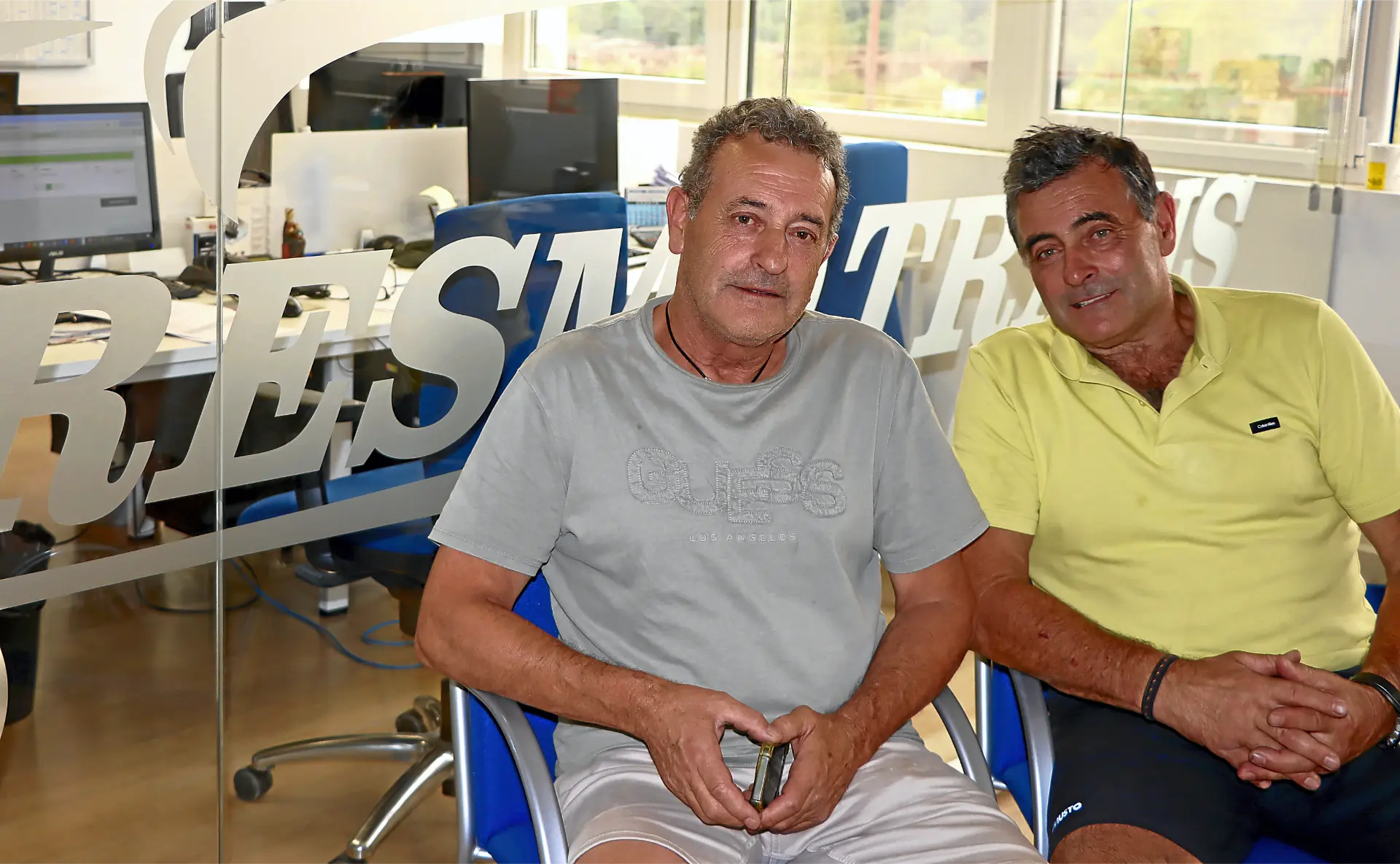 Dos hombres de mediana edad se sientan en sillas azules en una oficina con paredes de cristal y ordenadores al fondo, discutiendo sobre los camiones clásicos de Cresmotrans.