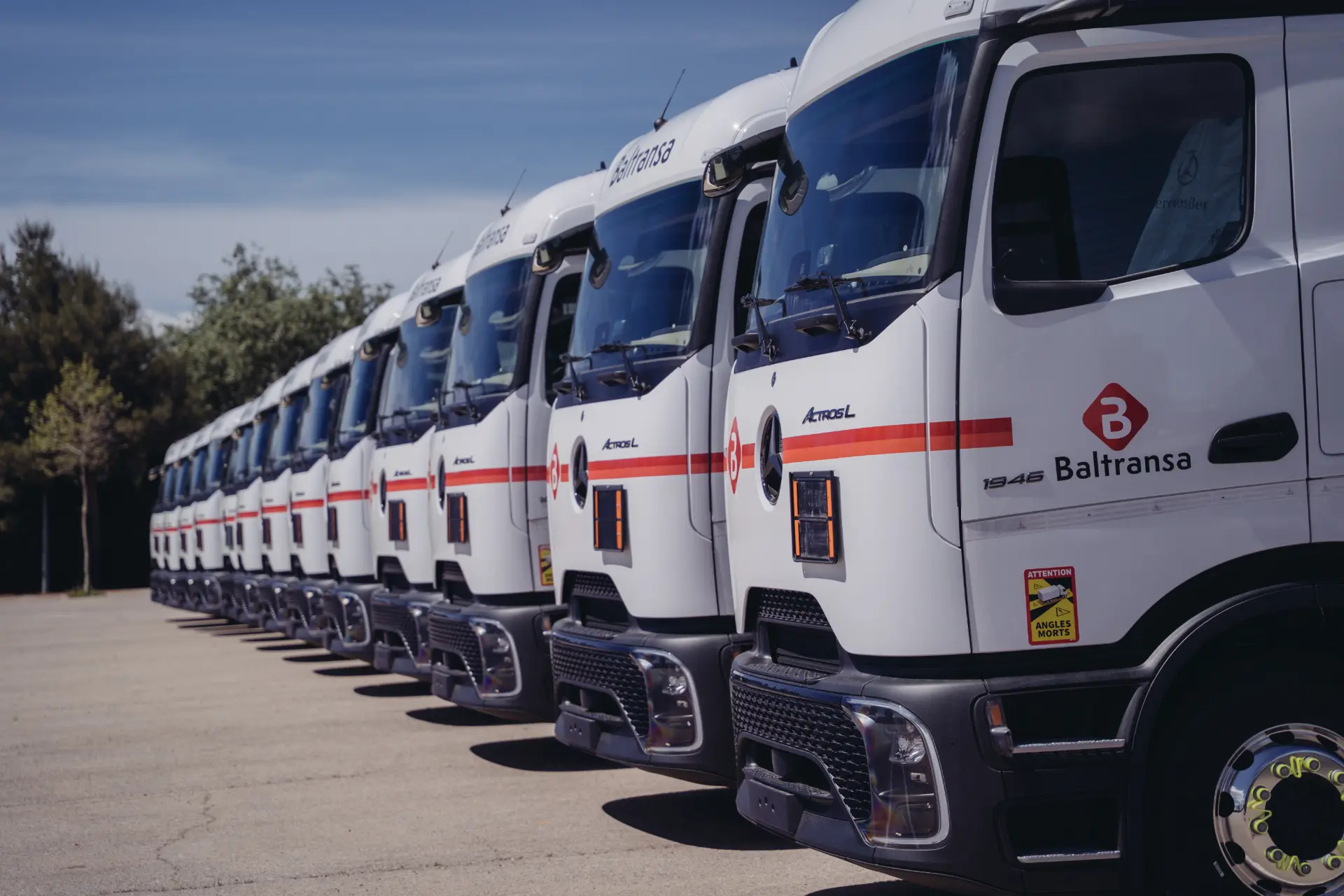 Una fila de camiones blancos de Baltransa, cada uno con el sistema de borrador automático de la empresa, aparcados uno junto al otro al aire libre en un día soleado.