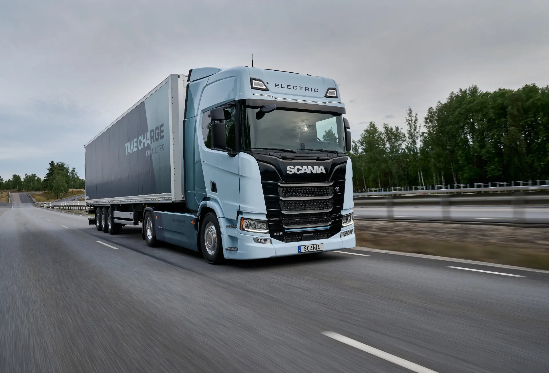Un semirremolque eléctrico Scania de color azul claro, que forma parte de la gama de camiones eléctricos y cuenta con el apoyo de Scania Services 360, circula por una autopista con árboles al fondo.