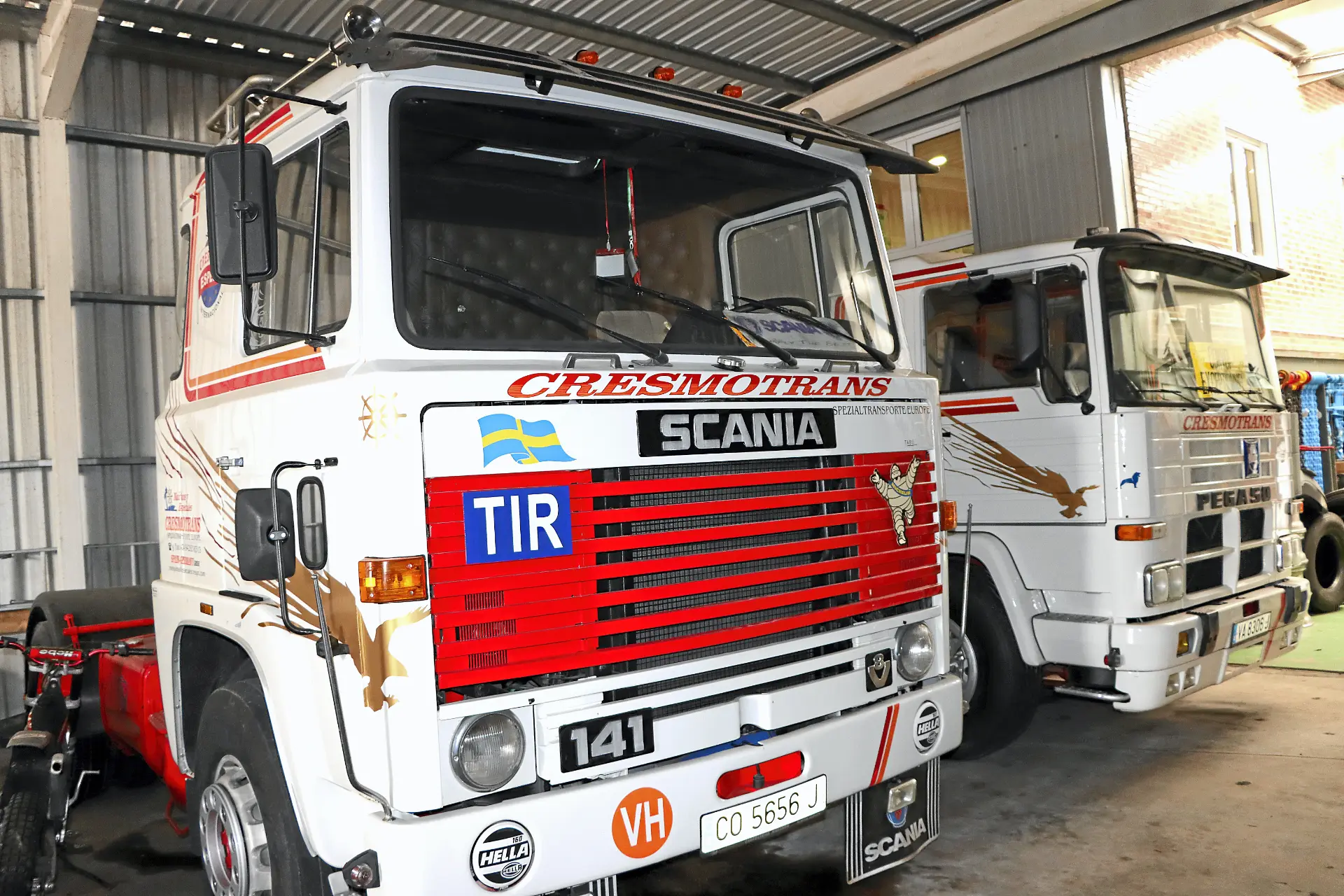 Dos camiones Scania clásicos blancos con detalles rojos, marcados con Cresmotrans, están aparcados en el interior de un garaje con techo metálico, mirando hacia delante, mostrando la calidad del transporte de Náuticos & Especiales.
