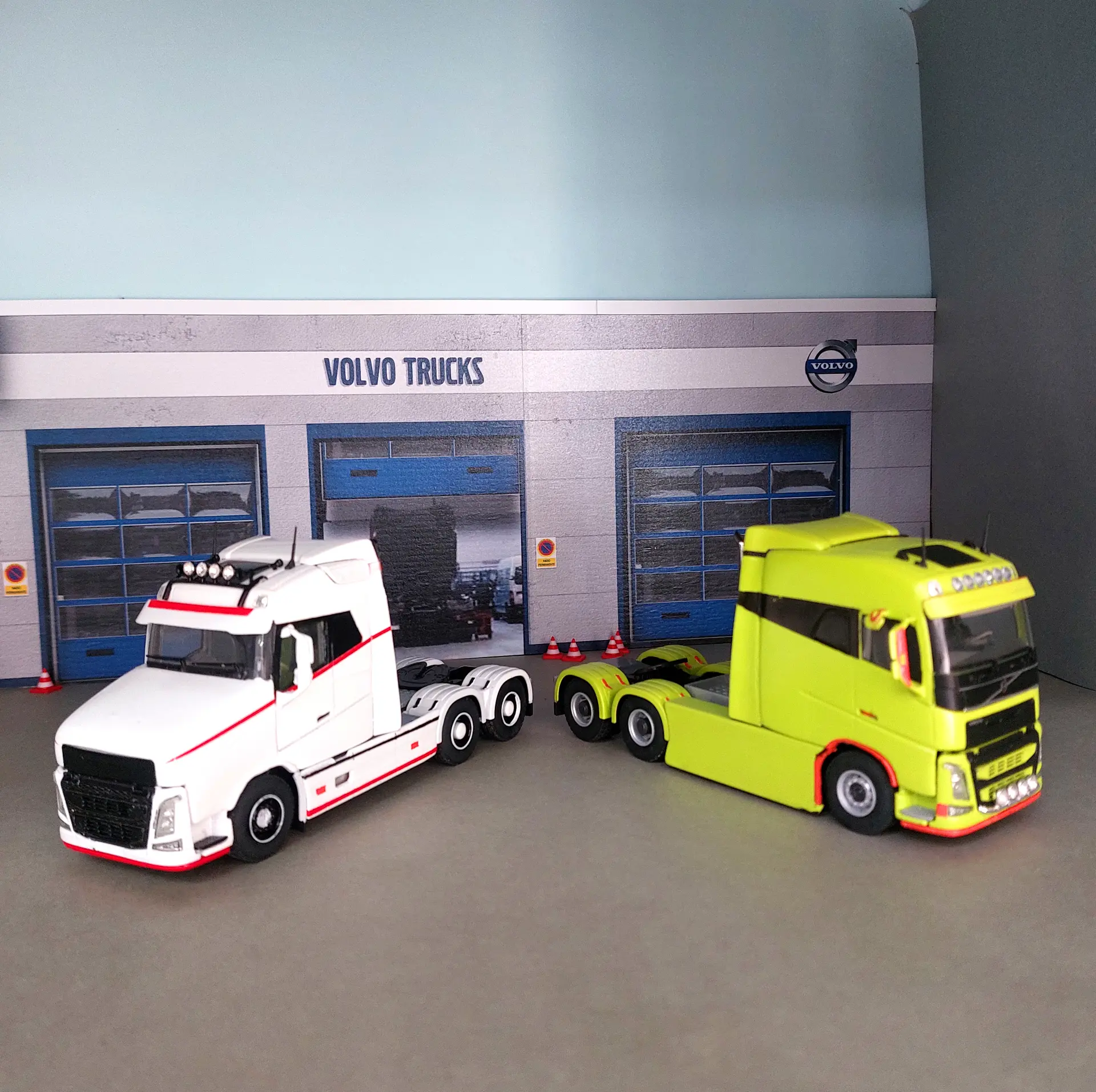 Dos modelos de camión Volvo, uno blanco con ribetes rojos y otro verde brillante, se sitúan frente a un fondo de garaje de camiones Volvo, perfecto para cualquier colección de Juan Carlos Carrasco o Siembre.