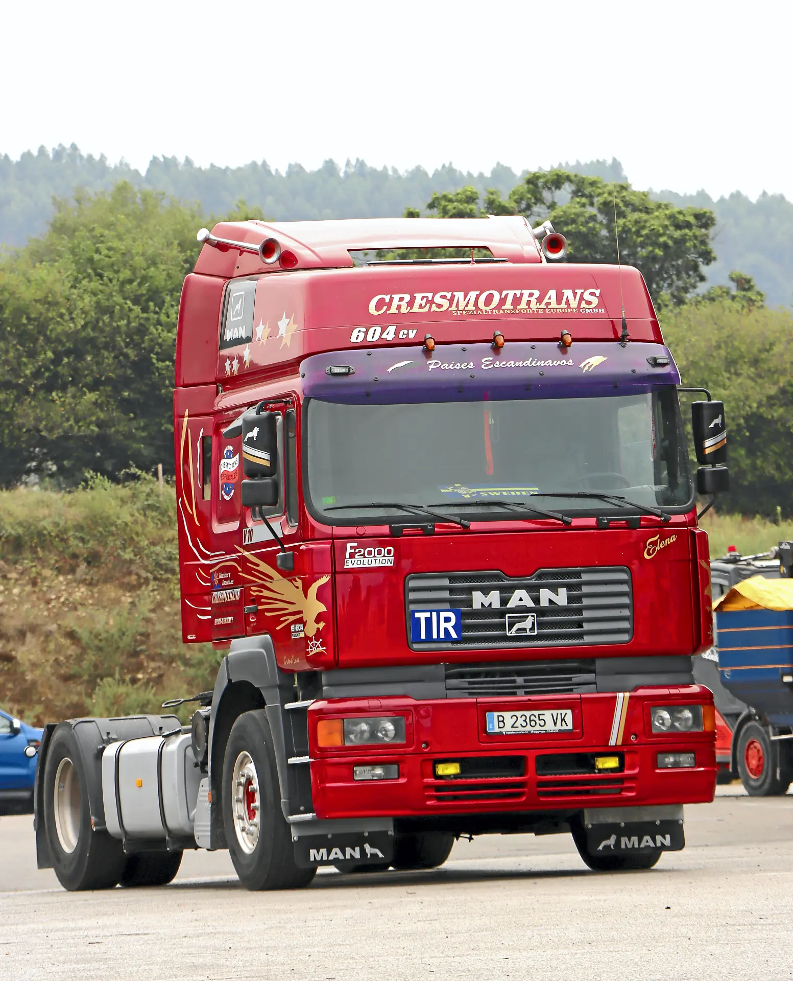 Camión MAN rojo con el logotipo "Cresmotrans" aparcado al aire libre, rodeado de árboles y otros vehículos, destaca entre los camiones clásicos.