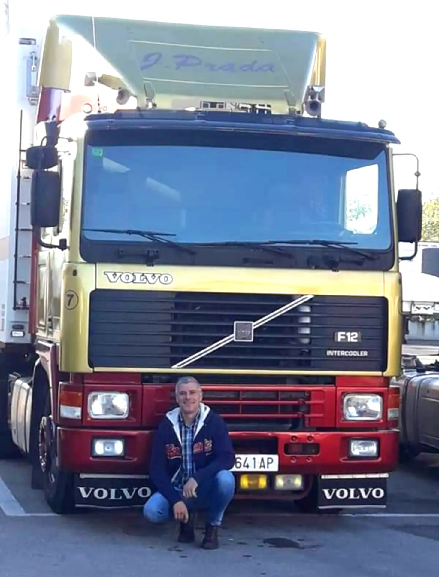 Juan Carlos Carrasco arrodillado y sonriente delante de un camión Volvo F12 amarillo y rojo aparcado en un solar, listo para otro día de siembre y recolecta.