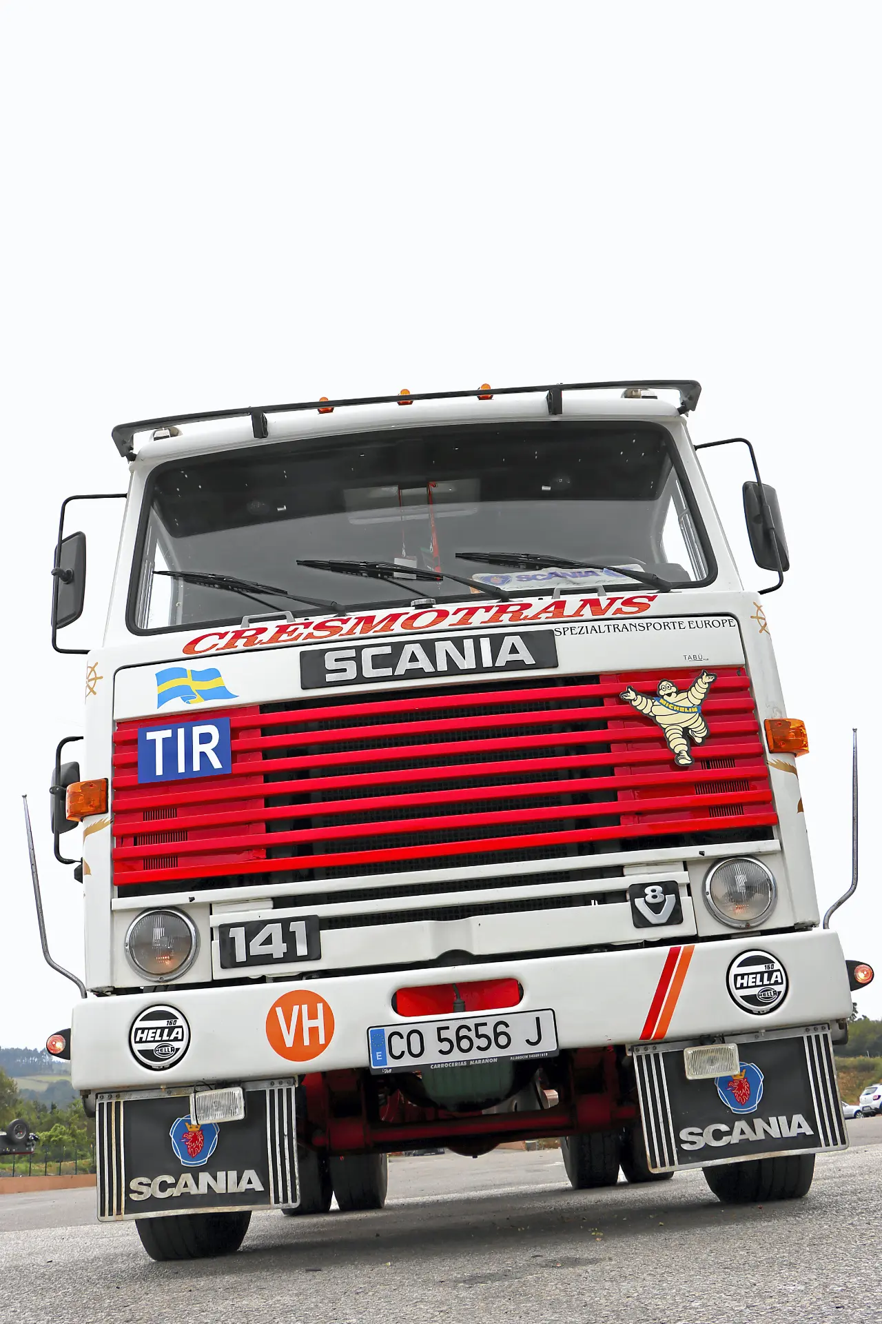 Camión Scania vintage blanco con rayas rojas y bandera sueca, aparcado en una carretera, vista frontal. Perfecto para aficionados a los camiones clásicos y coleccionistas en eventos Cresmotrans Náuticos & Especiales.