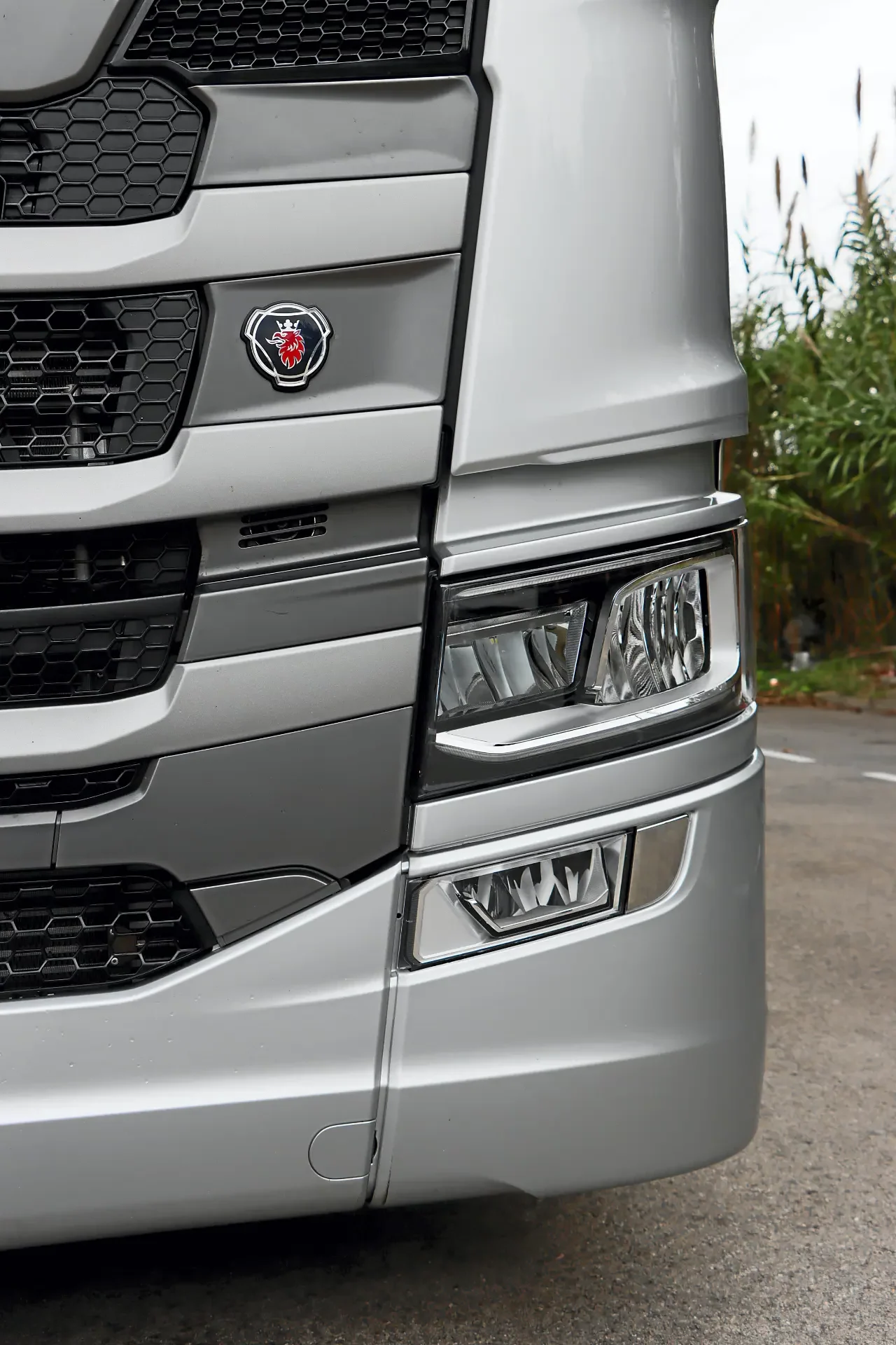 Primer plano de la parrilla delantera y los faros de un camión Scania plateado con borrador automático, aparcado cerca de la hierba.