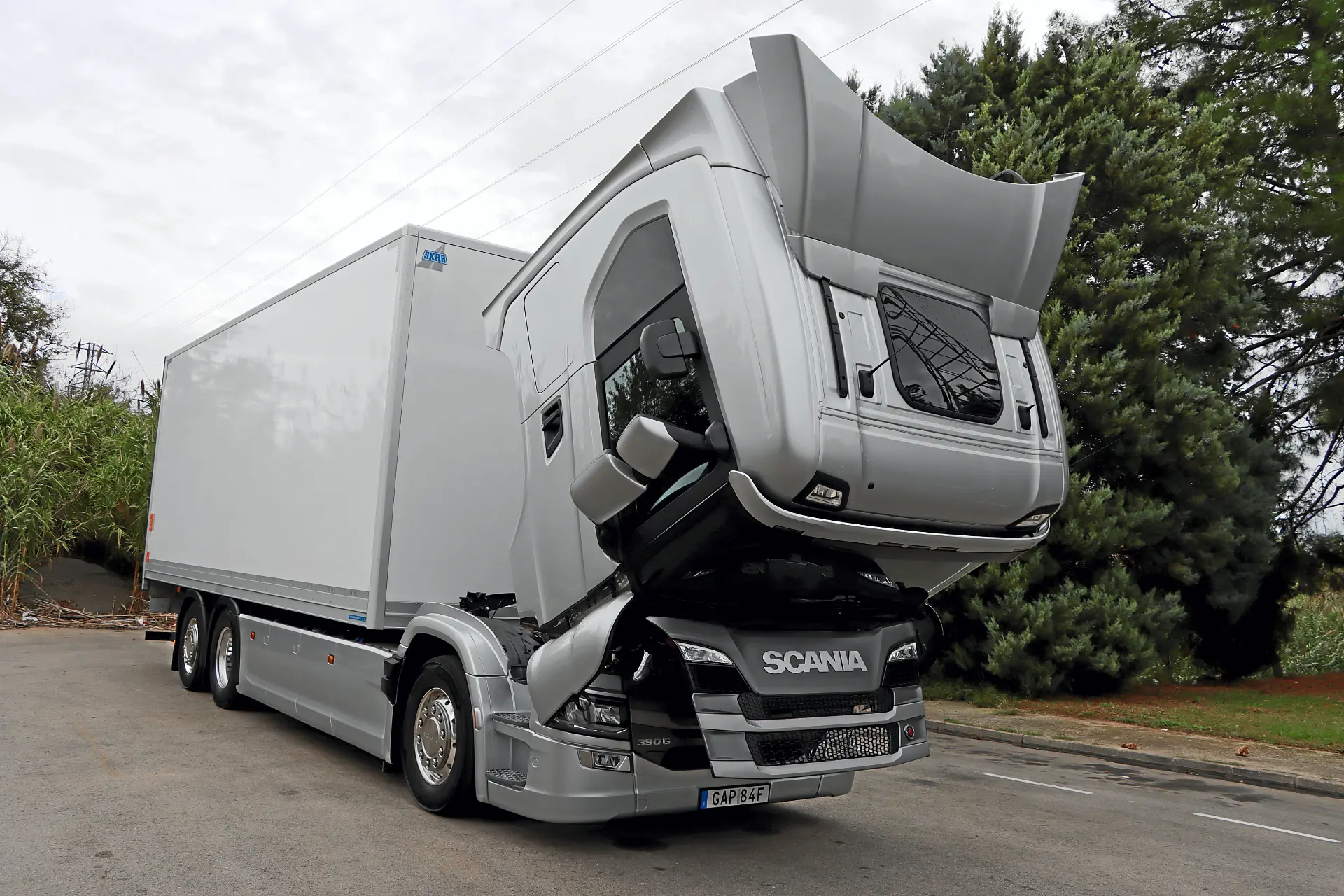 Un camión Scania plateado con la cabina inclinada hacia delante, dejando al descubierto el motor y aparcado en una carretera, parece listo para el mantenimiento o la inspección del borrador automático.