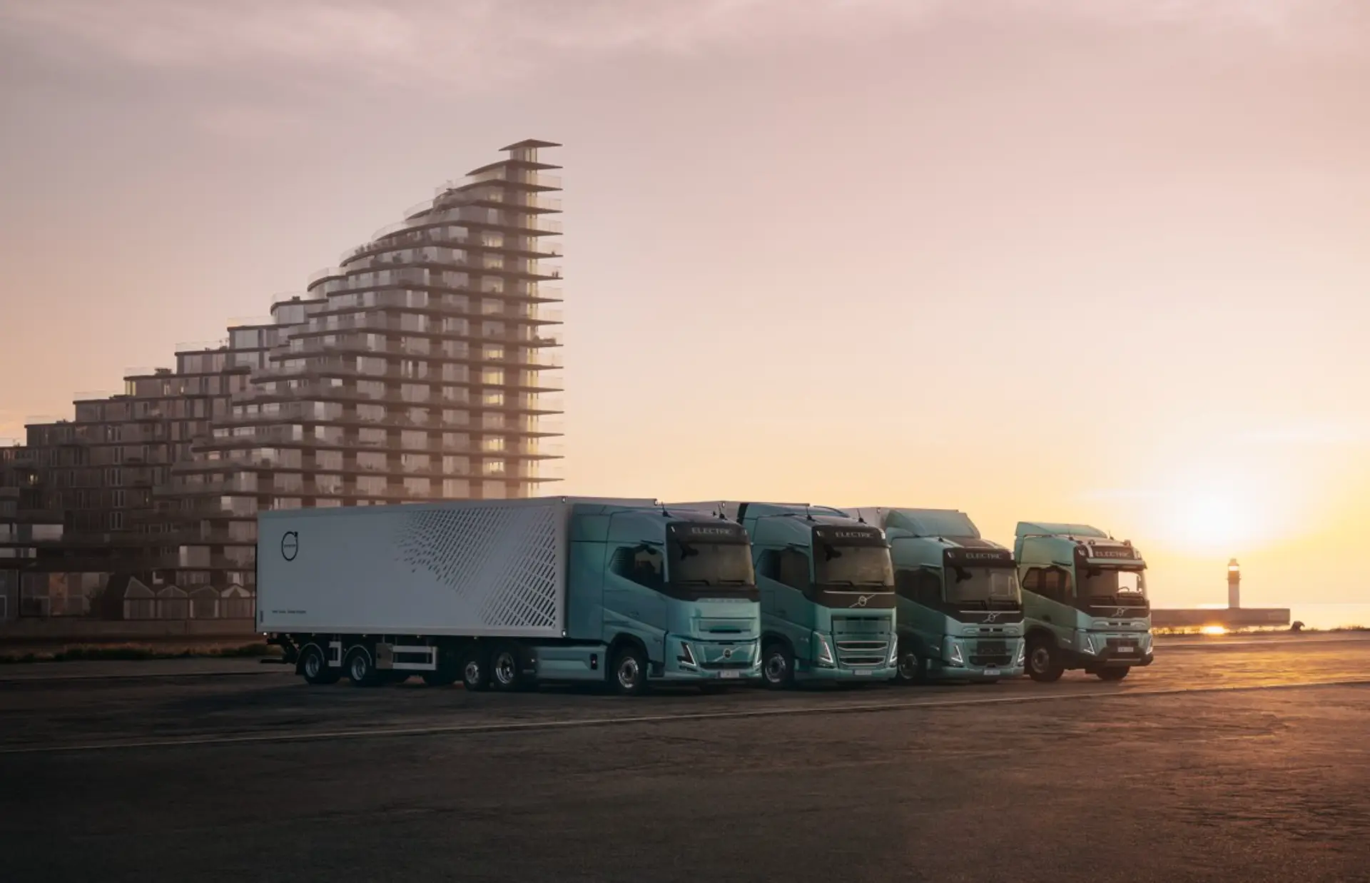 Cuatro semirremolques de color verde azulado, alineados uno al lado del otro cerca de modernos edificios al atardecer, esperan su próximo viaje, cada uno equipado con un borrador automático de última generación para realizar entregas eficientes.
