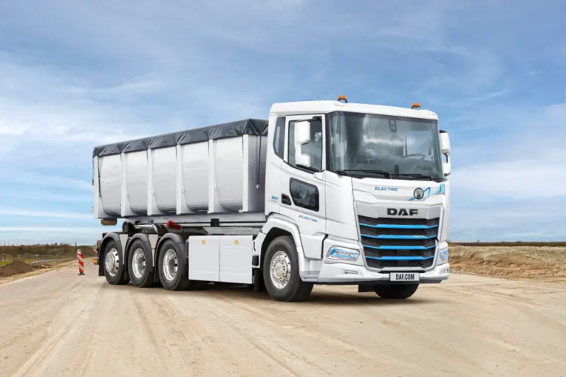 A white DAF electric dump truck, parte de los vehículos eléctricos y soluciones de carga modernas, está estacionado en un sitio de construcción arenoso bajo un cielo azul.
