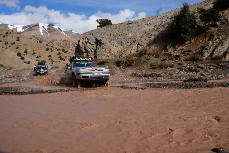 Dos todoterrenos se embarcan en una aventura 4x4, atravesando un río fangoso en un paisaje rocoso y montañoso bajo un cielo parcialmente nublado, capturando el espíritu del próximo Volkswagen Golf Challenge.