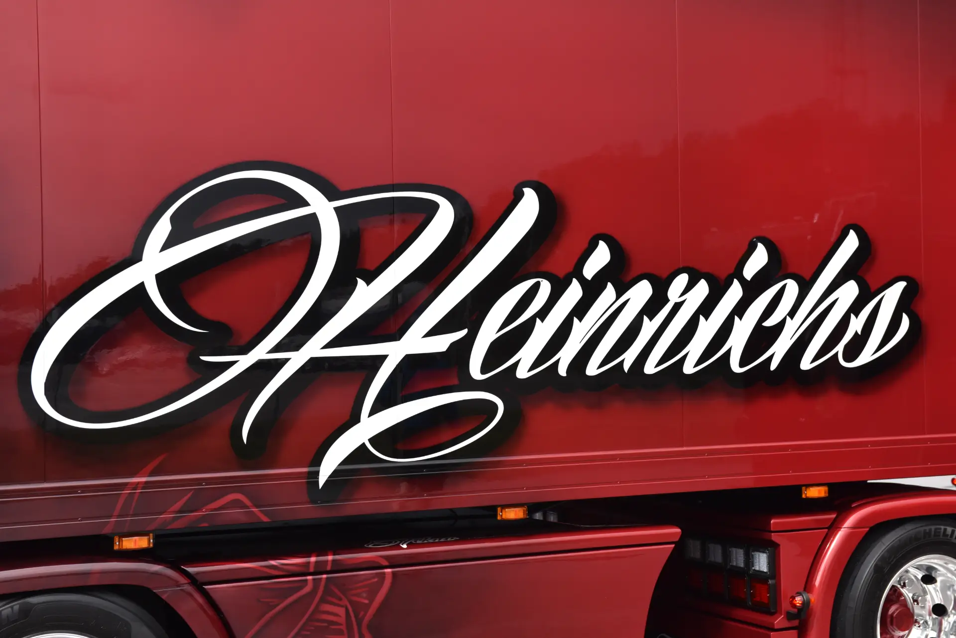 El lateral de un camión Scania R770 rojo muestra la palabra "Heinrichs" en grandes letras cursivas blancas, reflejo de la pasión que Carmen y Sascha Heinrichs ponen en su trabajo.