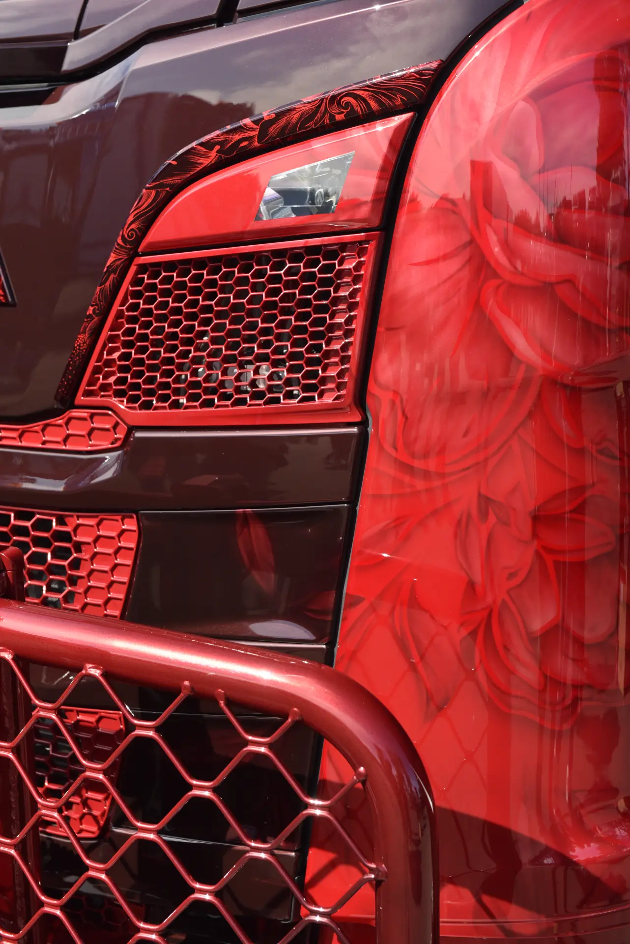 Primer plano del frontal pintado en rojo y negro de un camión Scania R770 con motivos florales, una rejilla de malla metálica y un toque de pasión en el detallado diseño.