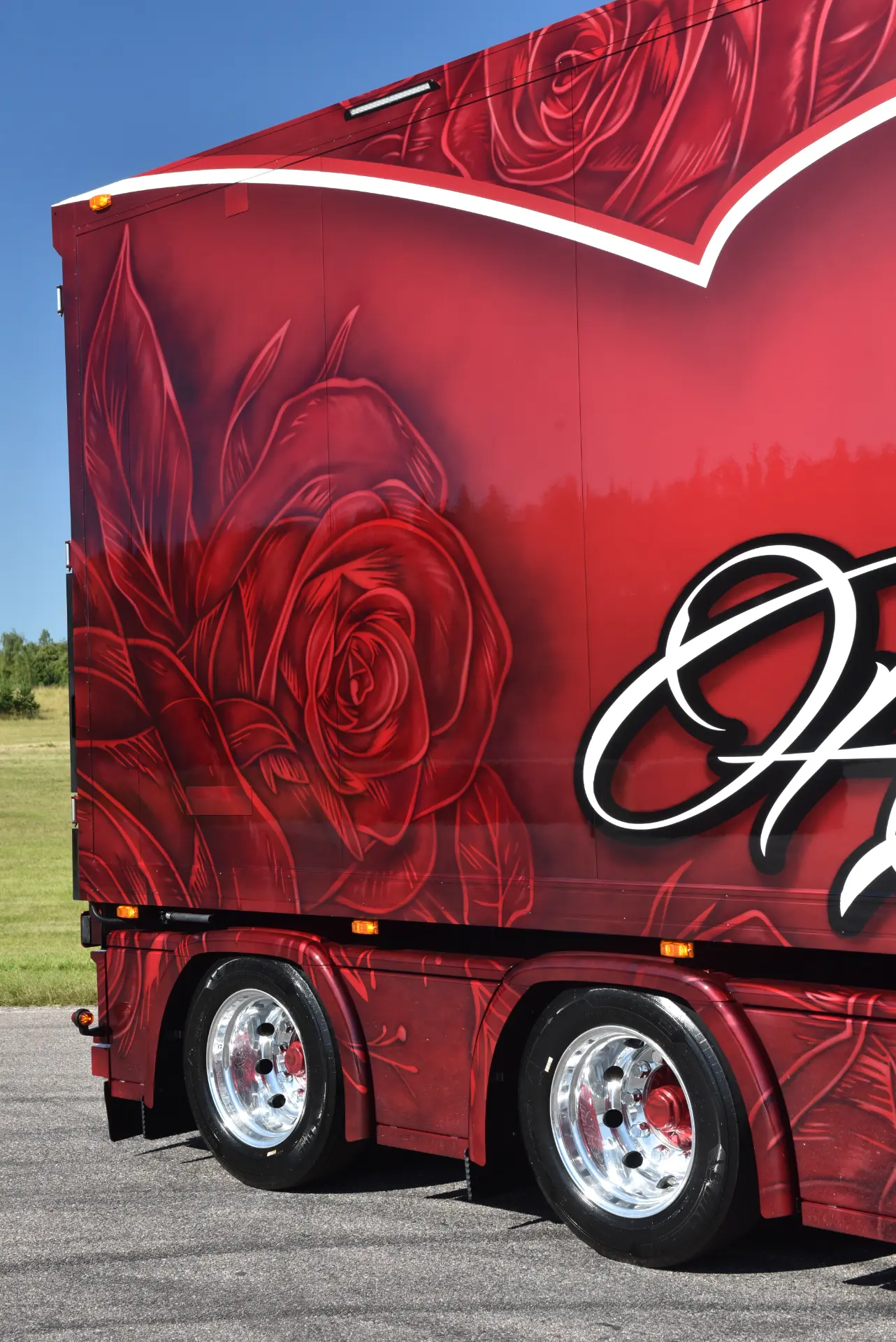 Remolque de camión Scania R770 rojo con grandes y detalladas ilustraciones de rosas que irradian pasión, llantas cromadas relucientes: un llamativo homenaje de Carmen y Sascha Heinrichs aparcado en la carretera.
