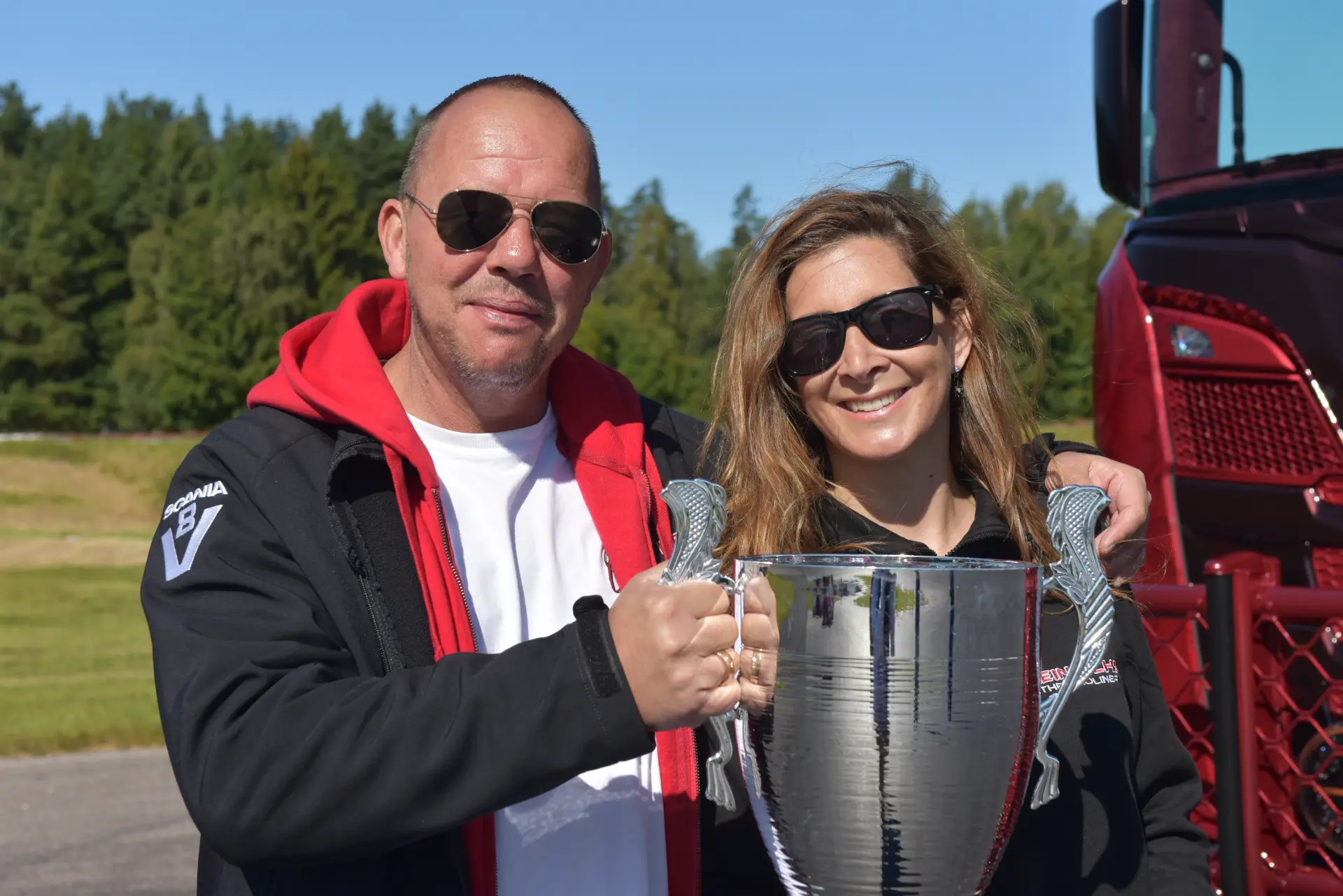 Carmen y Sascha Heinrichs, con gafas de sol, sonríen y sostienen un gran trofeo al aire libre en un día soleado, celebrando su pasión por el éxito.