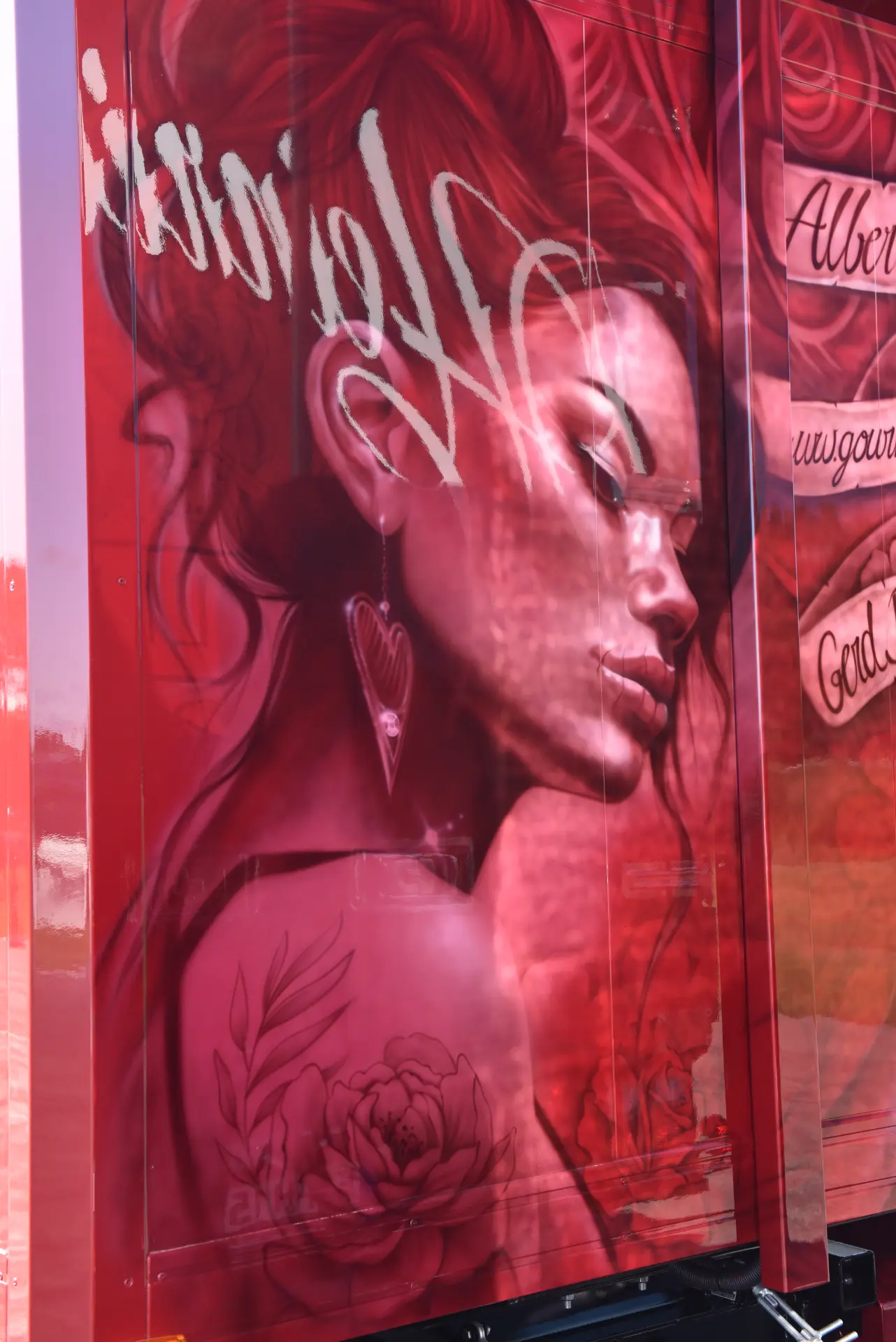Mural aerografiado de una mujer con tatuajes florales, pendiente de corazón y pelo alborotado en tonos rojos y rosas, eco de la pasión que se ve en las vibrantes obras de Carmen y Sascha Heinrichs.