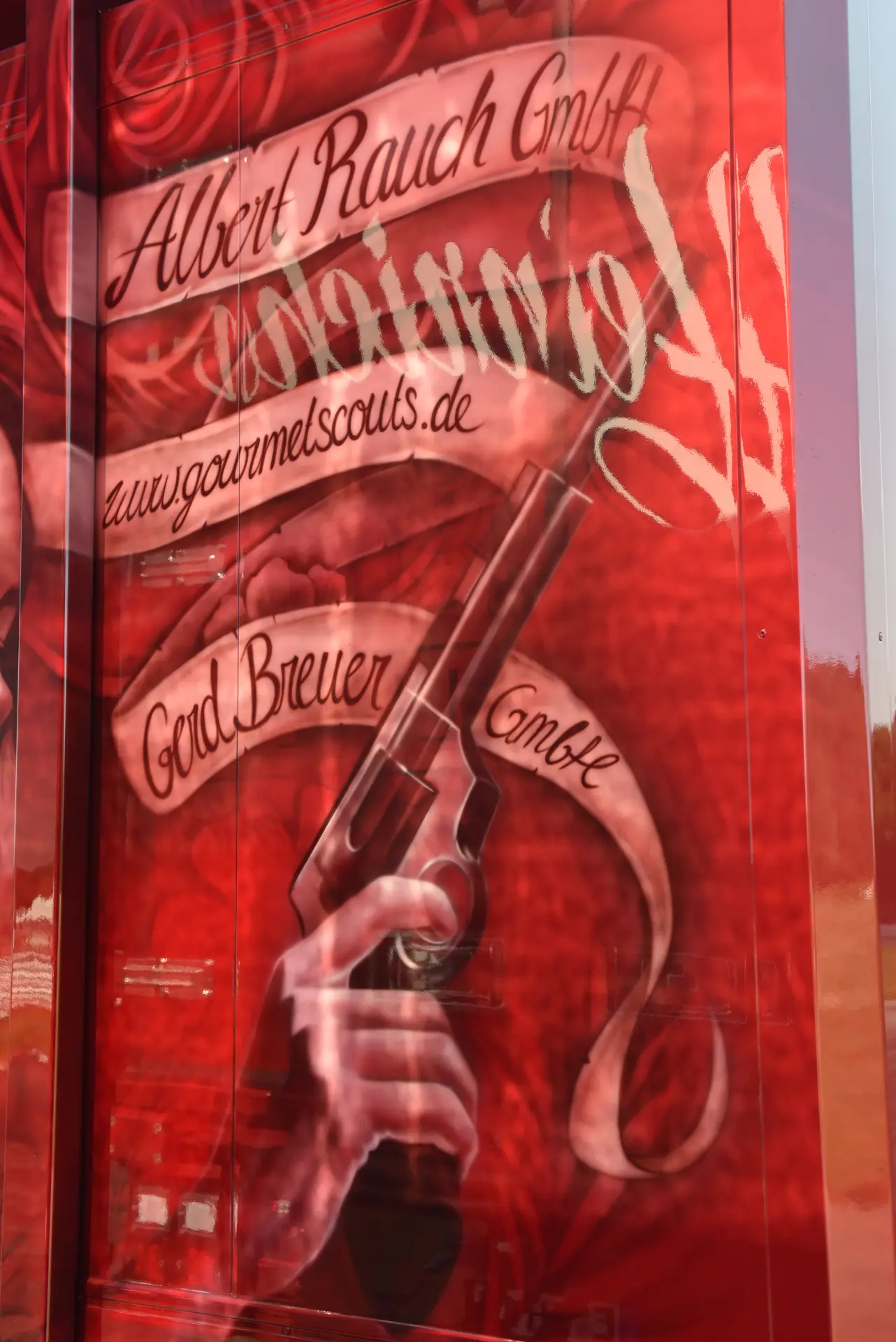 Mural rojo con una mano empuñando un revólver, rosas y texto en alemán con los nombres de la empresa y el sitio web. Incluye "Carmen y Sascha Heinrichs" junto al atrevido diseño.