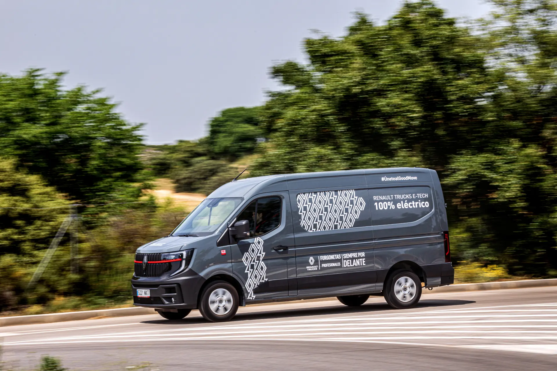 Una furgoneta de reparto eléctrica Renault Trucks Master gris circula por una carretera curva, rodeada de árboles y vegetación, mostrando la eficacia de las furgonetas eléctricas en el transporte moderno.